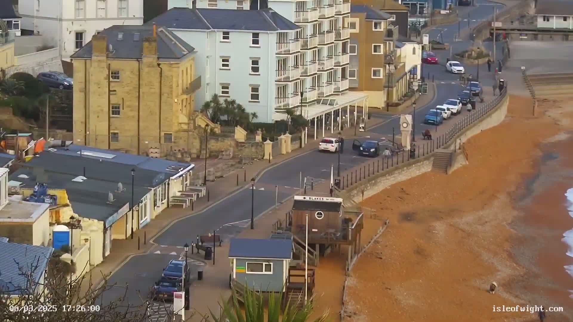 A seaside town street scene with buildings, parked cars, and a sandy beach, on an overcast day.