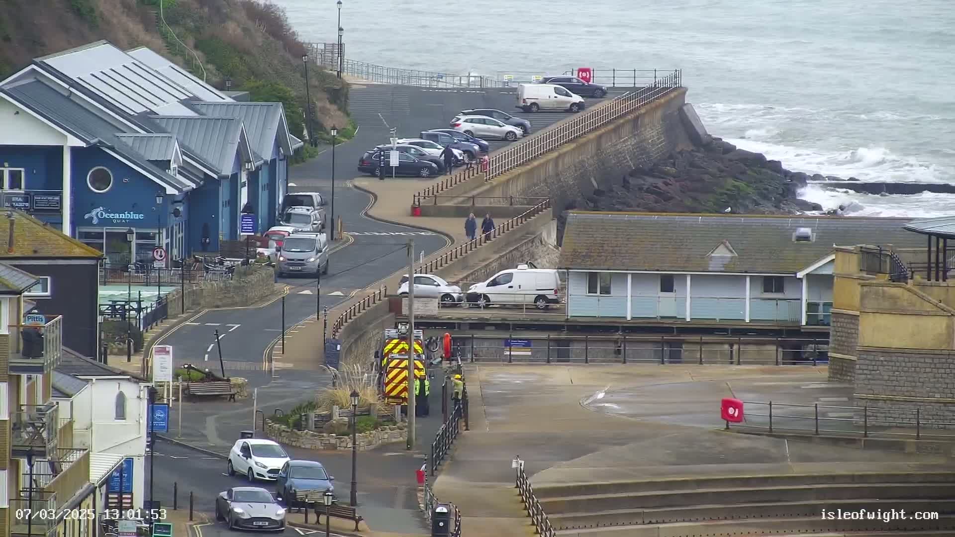 A coastal scene on an overcast day shows a road curving alongside a seawall with parked cars, buildings, and a few pedestrians; choppy waves crash against rocks below the wall.