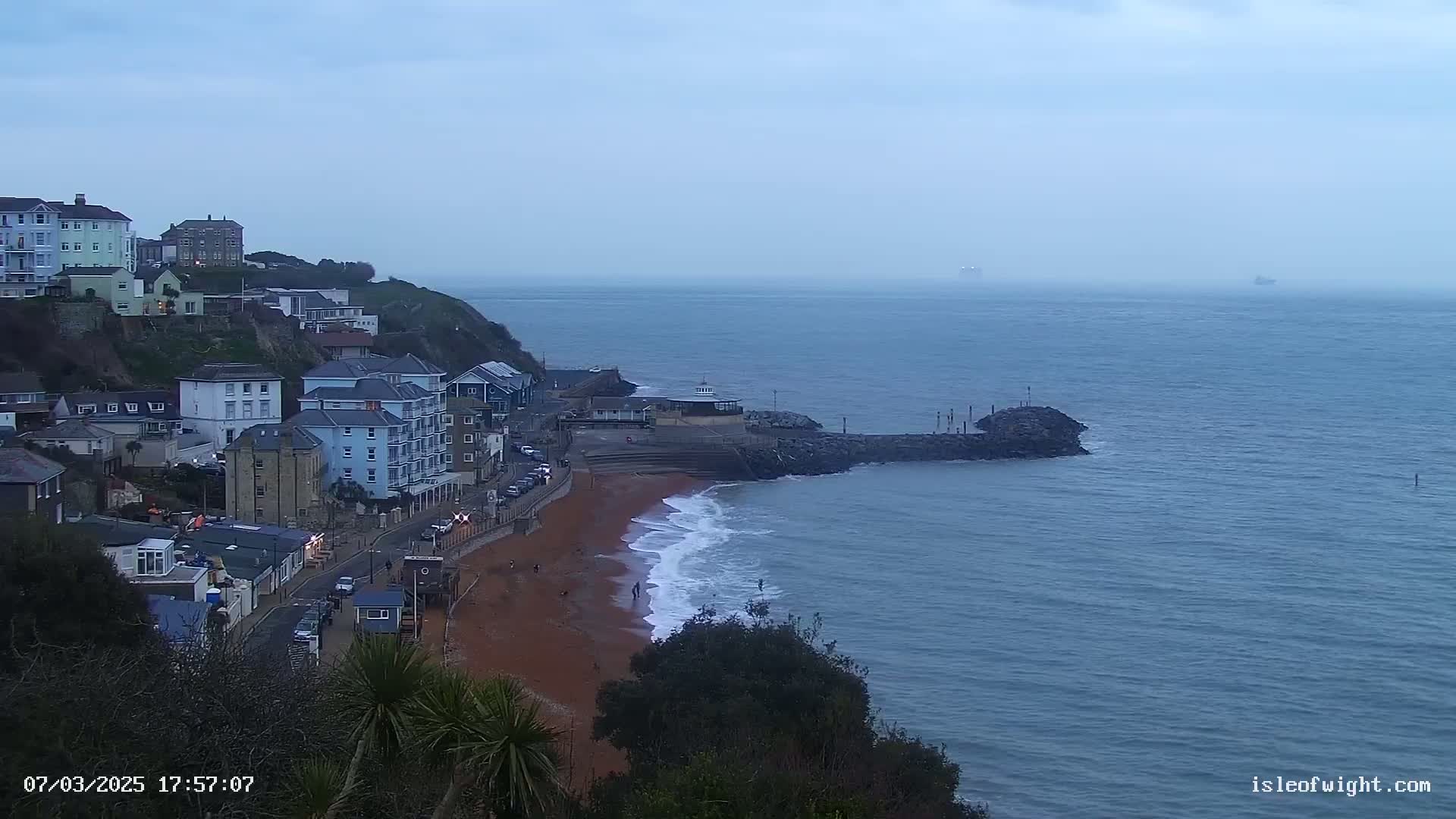 A red sand beach with small waves laps at a rocky breakwater, in front of a cliffside town under an overcast sky.