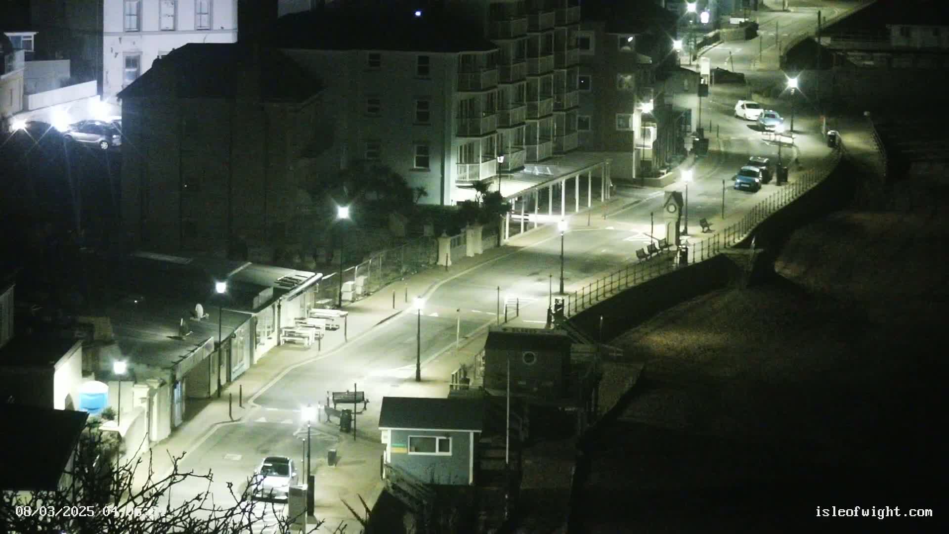 A nighttime, brightly lit street scene shows a road winding past buildings and a beach.
