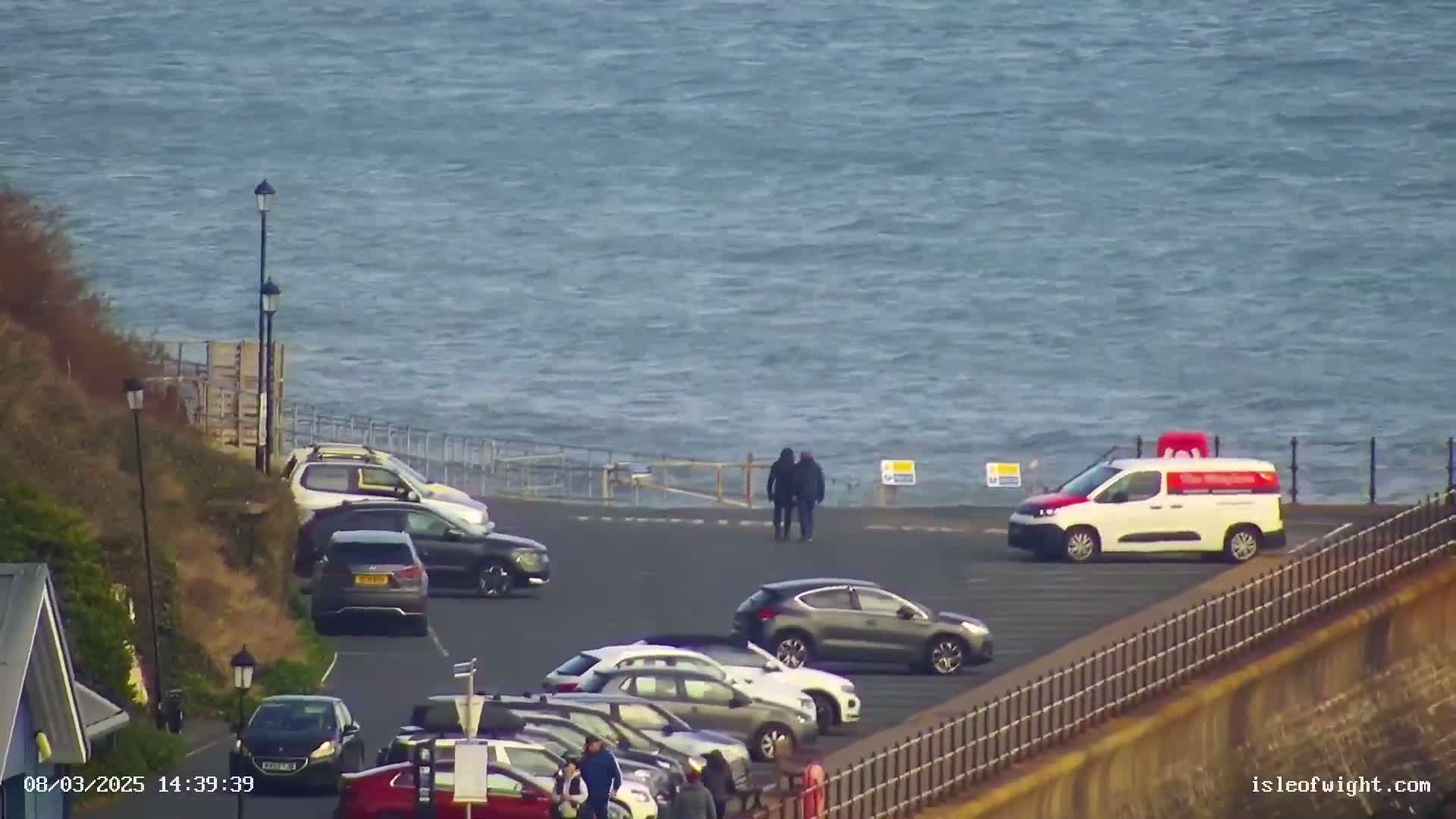 A parking lot overlooking a choppy sea is populated by several cars and two pedestrians, with a van parked nearby.