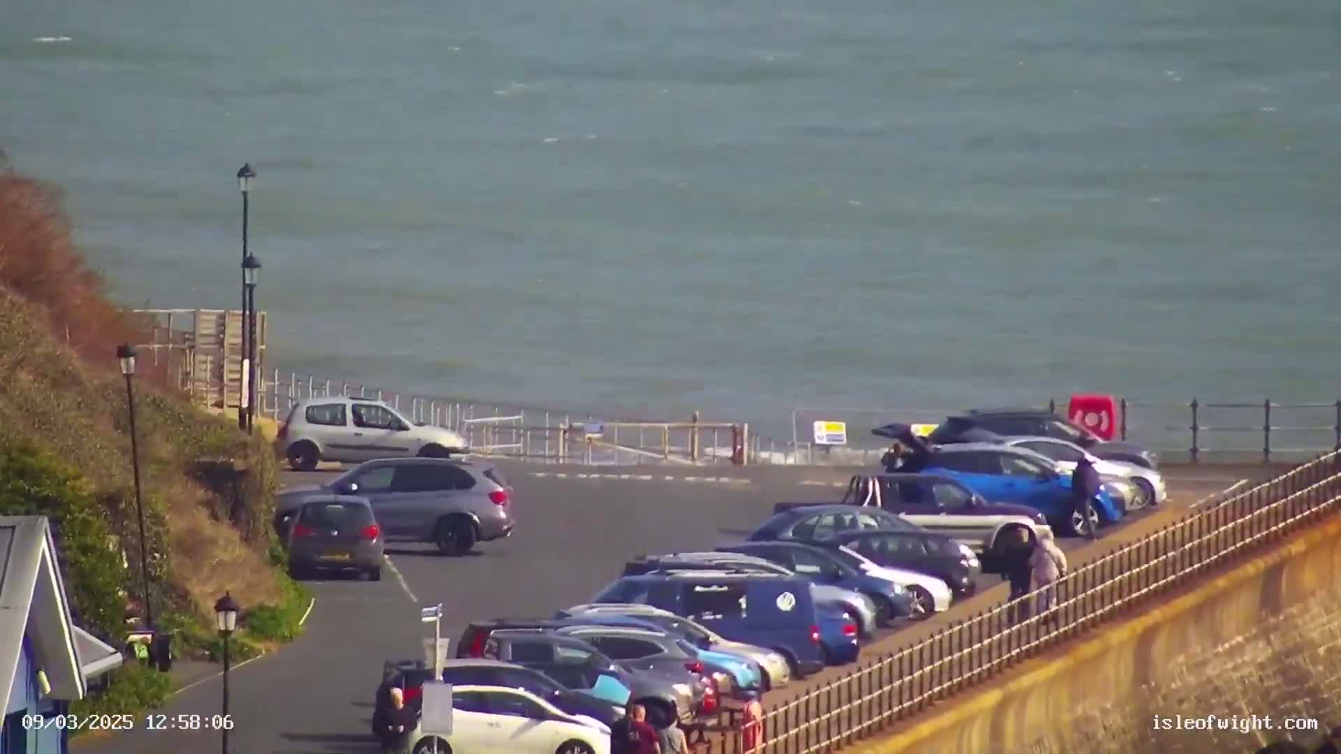 A parking lot overlooks a choppy sea on a sunny day, with several cars parked and a few people walking nearby.
