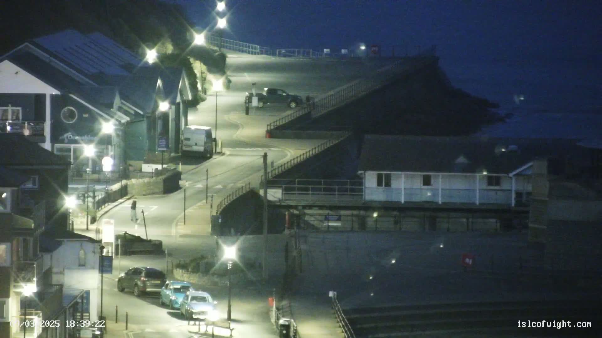 A nighttime coastal scene shows a curving road illuminated by streetlights, several parked cars, buildings, and a dark ocean in the background.