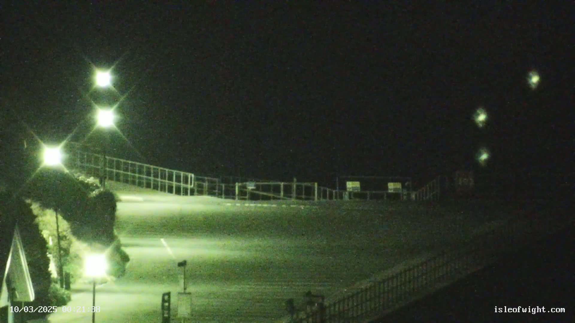 A nighttime view of an empty paved area, illuminated by several lights, with a fence and some vegetation visible.