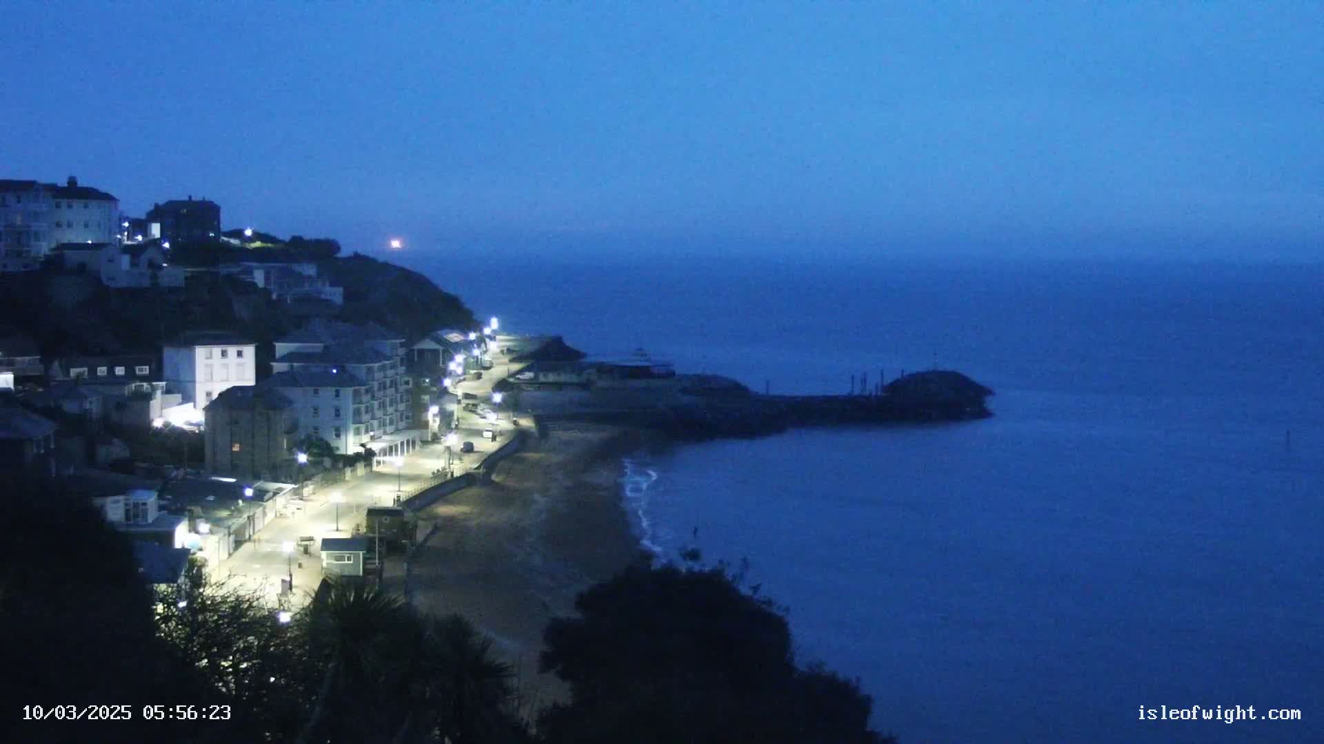 A seaside town at twilight, with lights illuminating a coastal path and beach, under a dark blue sky.