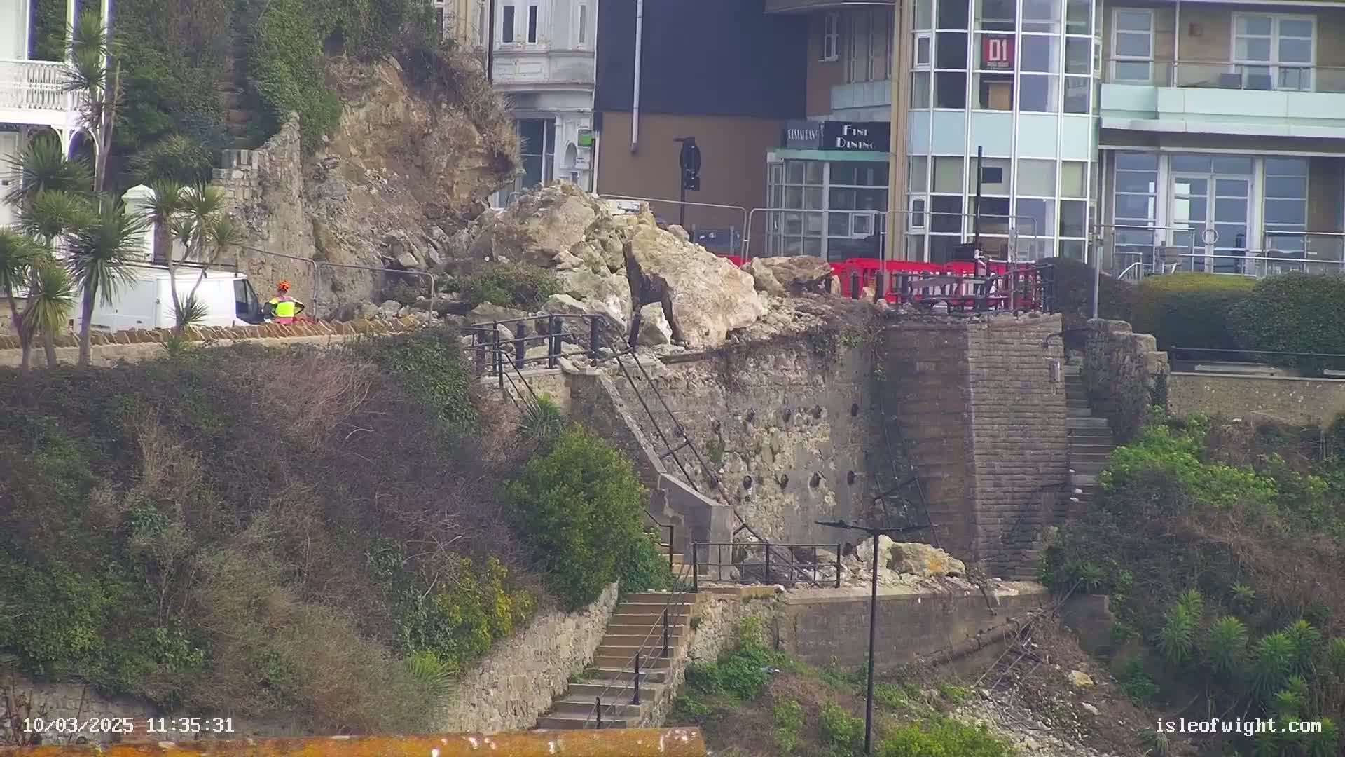 A section of a stone retaining wall has collapsed onto a set of stairs near a building, with a person in a safety vest visible near a van.