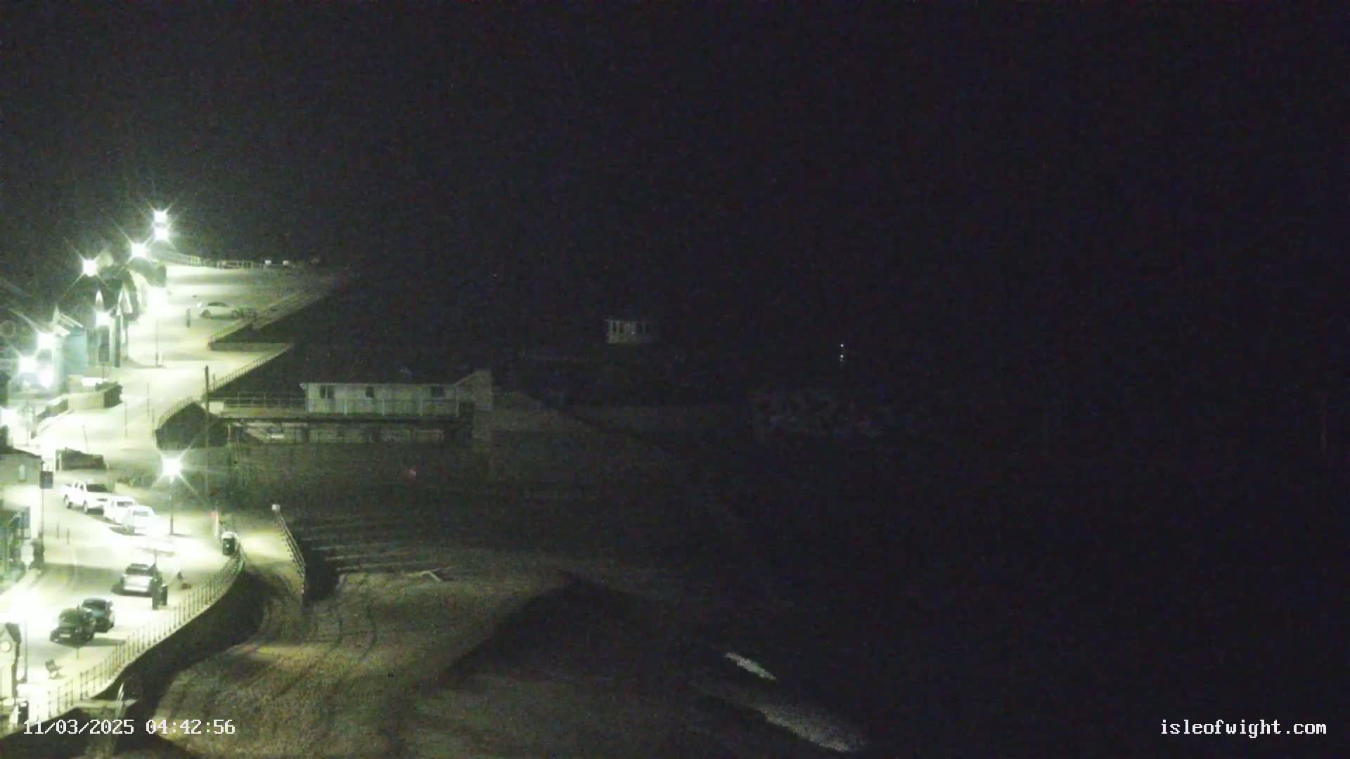 A nighttime view of a coastal road with parked cars, illuminated by streetlights, overlooking a dark ocean.