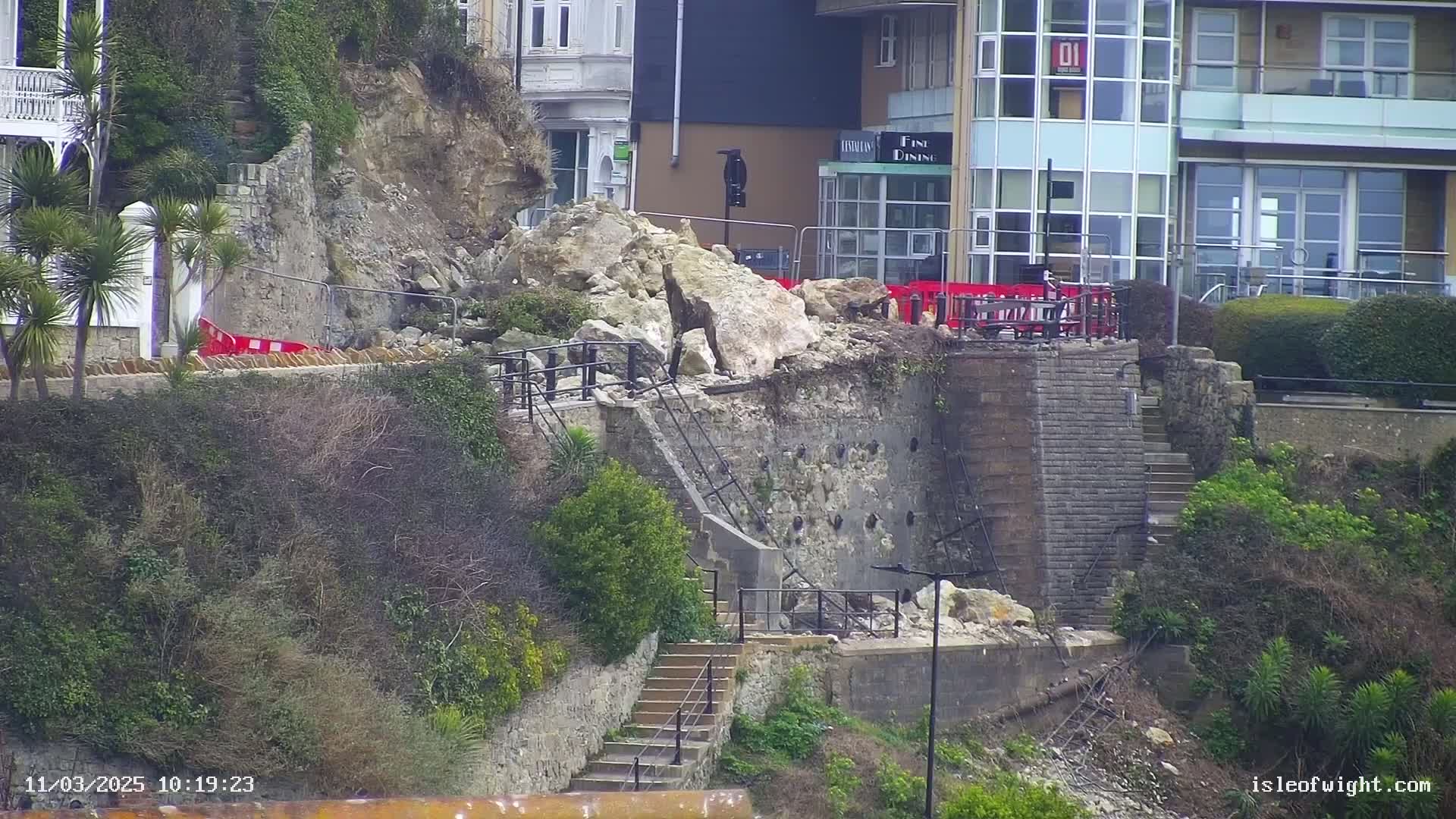 A section of a stone retaining wall has collapsed onto a set of stairs leading down a cliffside, near buildings on an overcast day.