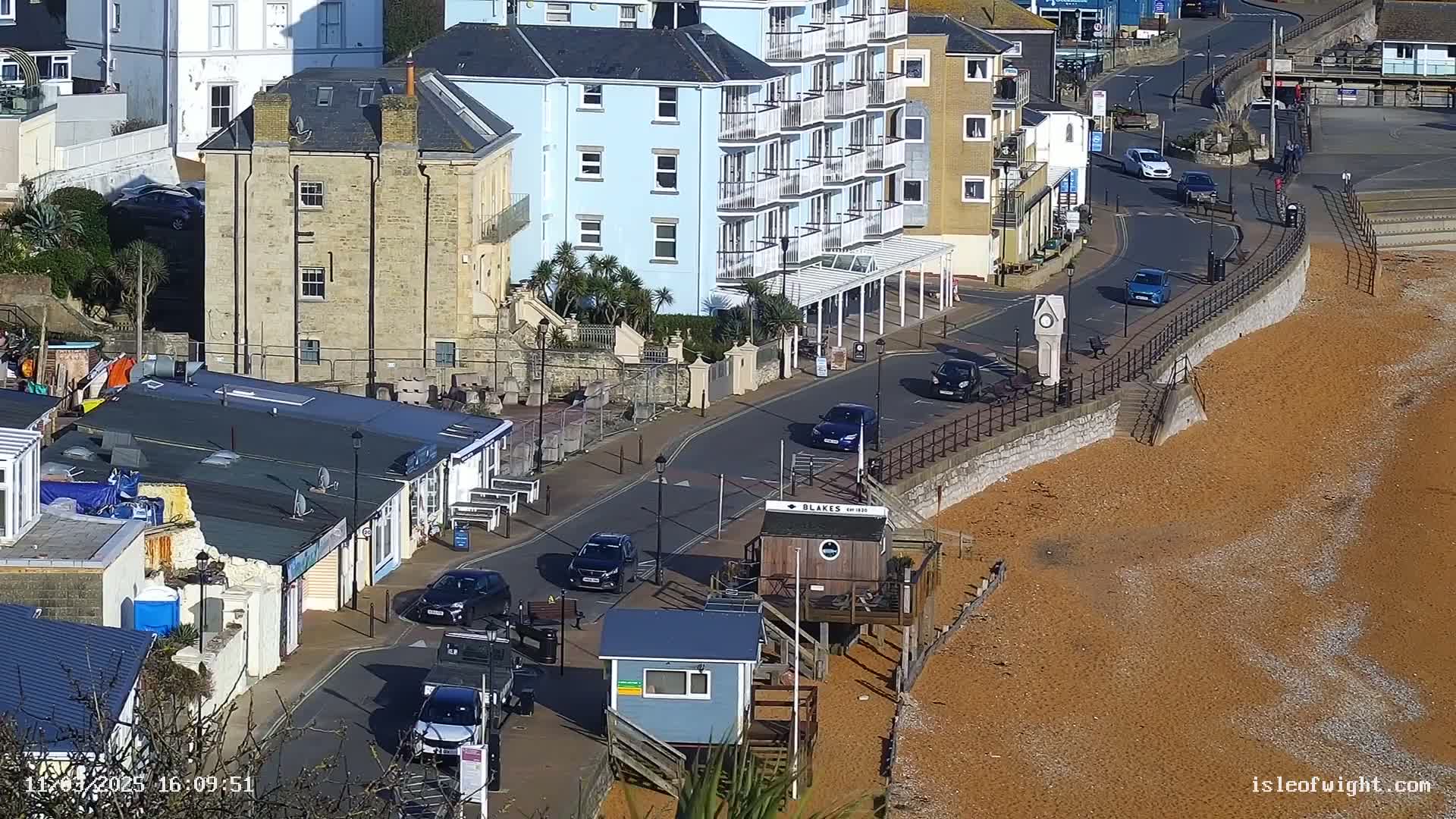 A sunny seaside town scene shows a curving road with several cars, buildings, and a beach with reddish-brown pebbles.