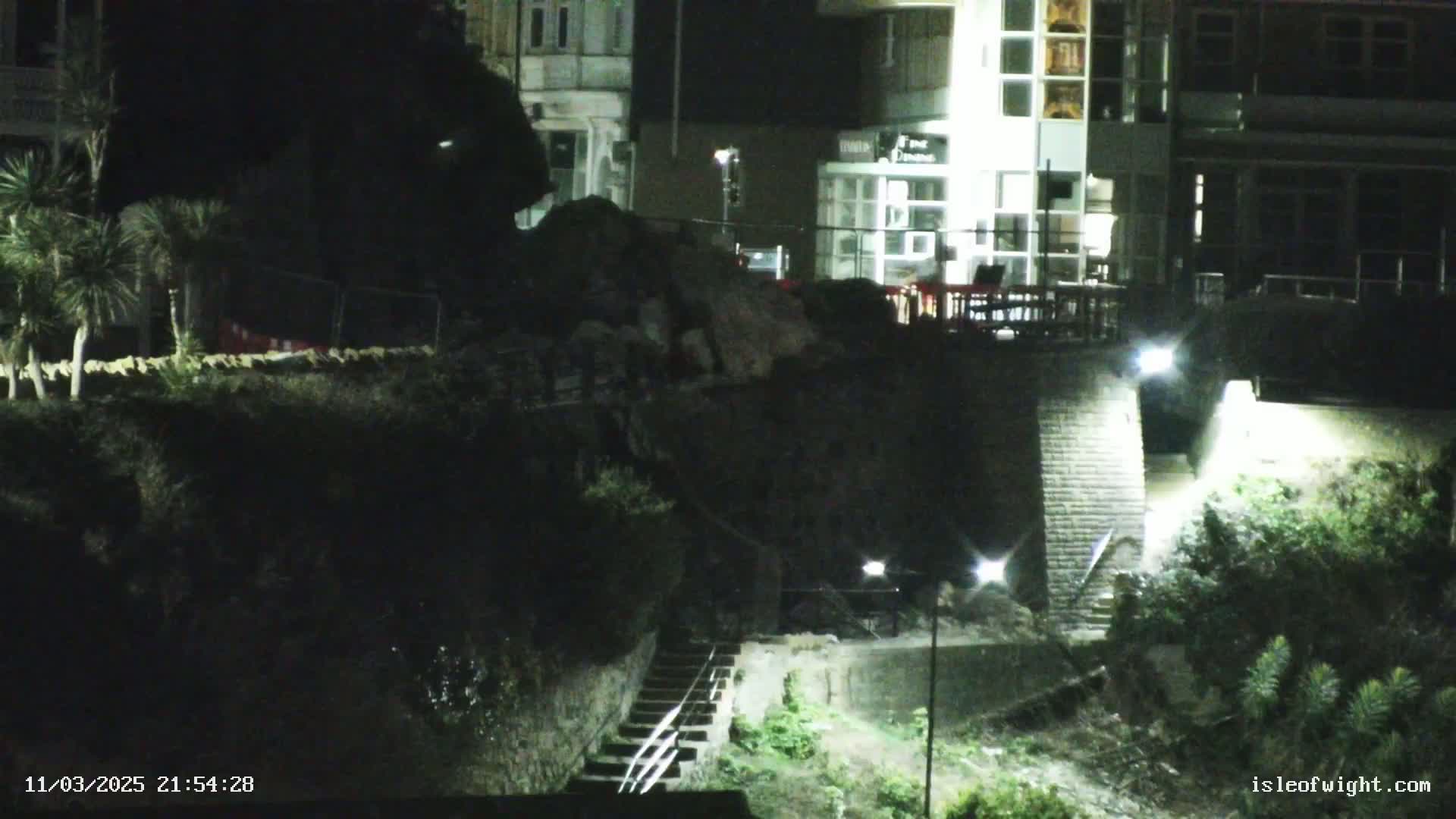 At night, a pile of rubble sits beside a lit stairway leading down to a dimly lit area below a building.