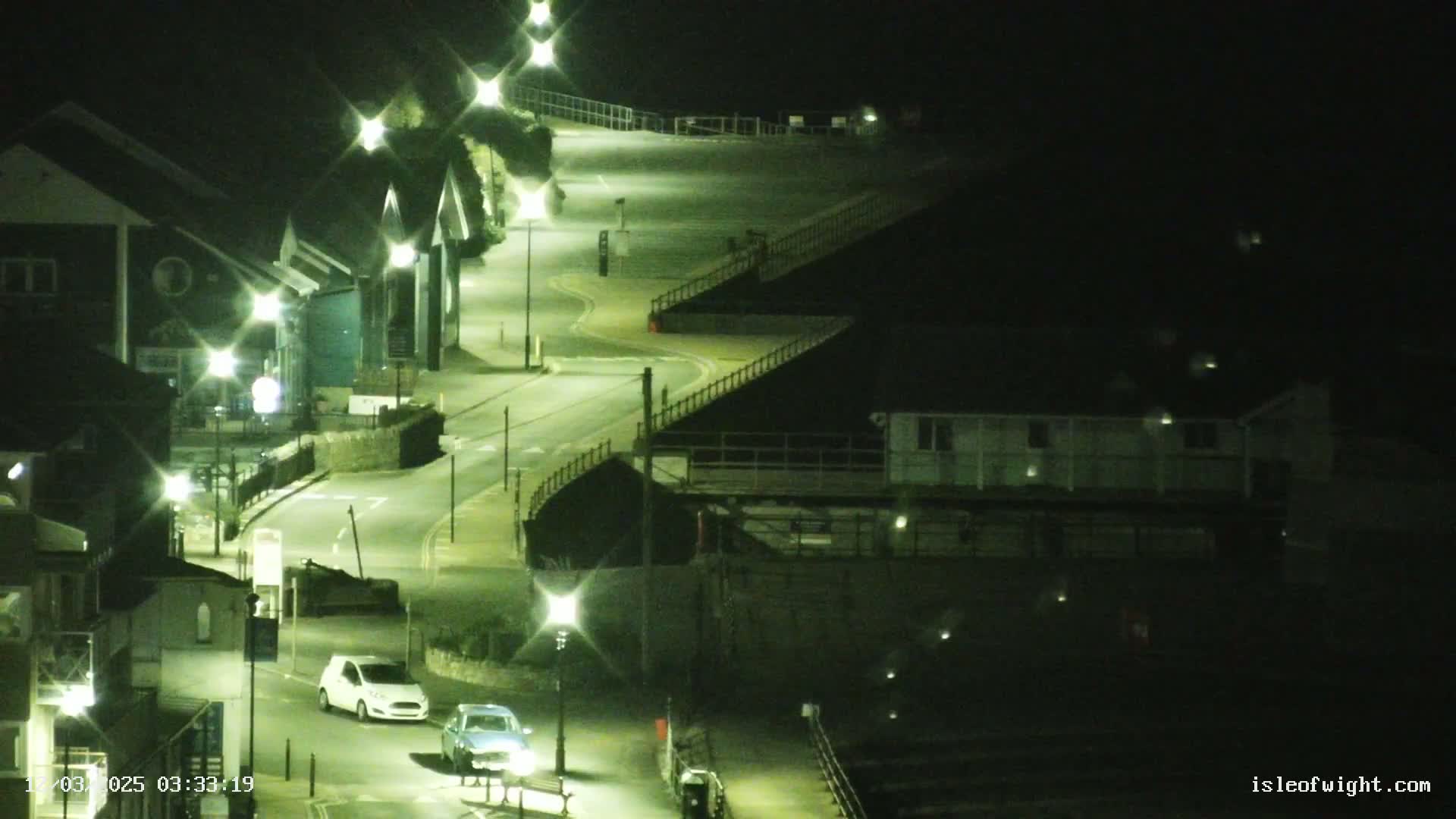 A winding road at night, illuminated by streetlights, passes by buildings and a sloped area with railings.