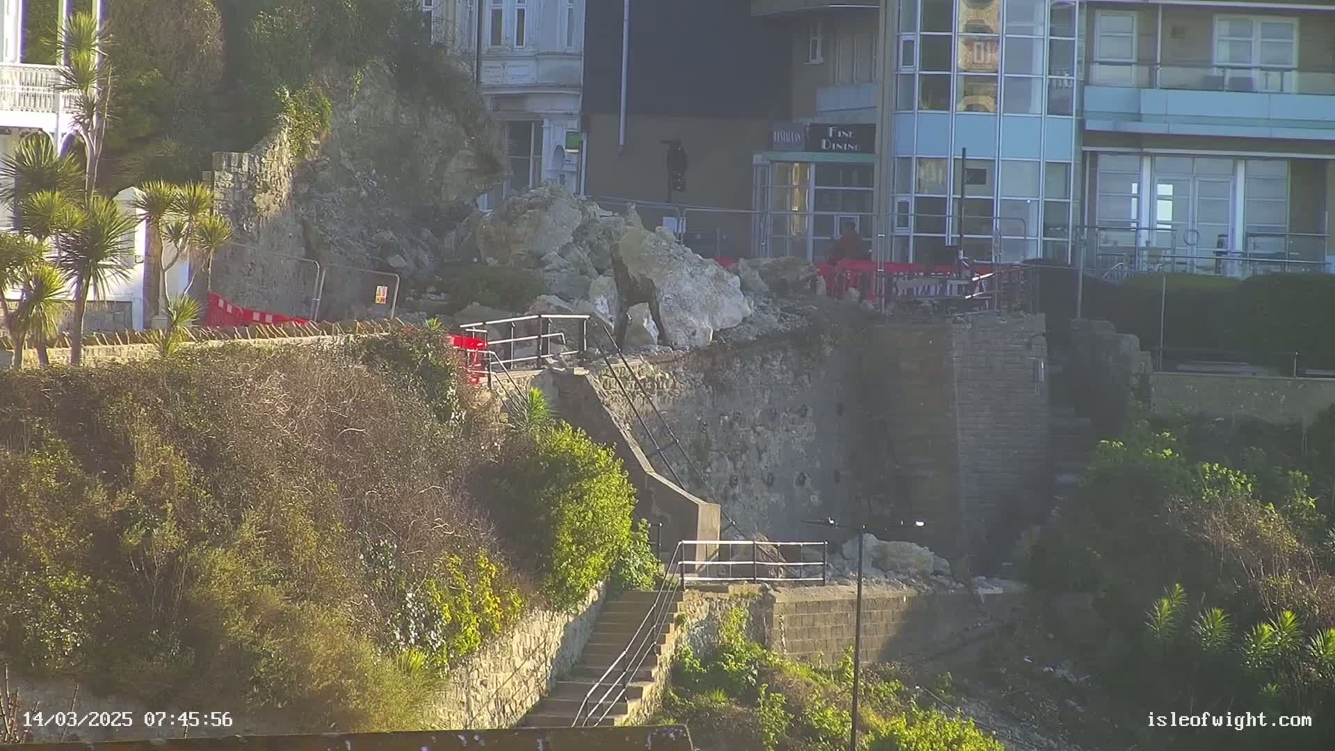 A stone retaining wall has collapsed onto a stairway leading down to the water, with large rocks and debris scattered below, near buildings in sunny conditions.