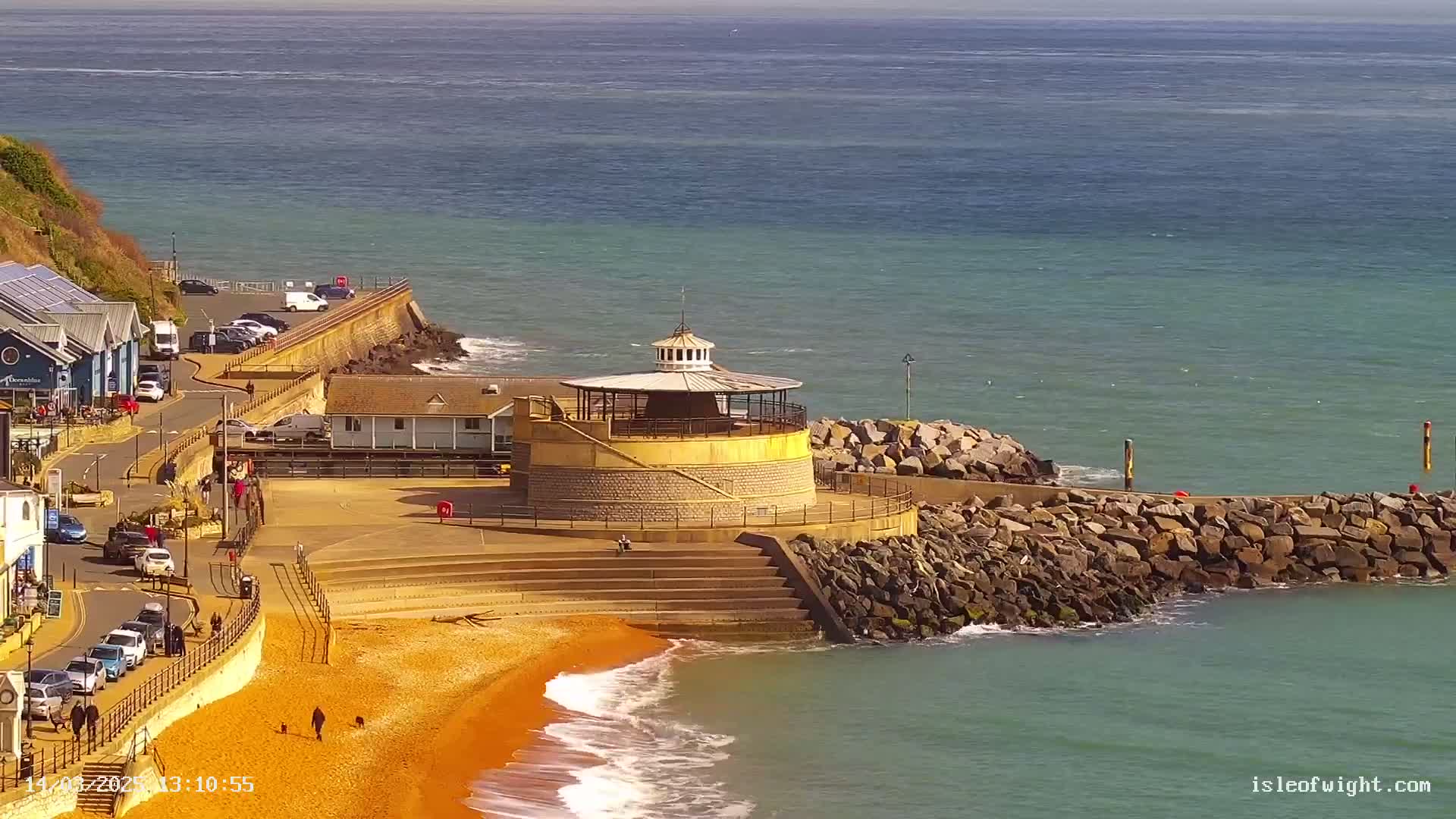 A coastal scene on a sunny day shows a round, tan-colored building with a white cupola on a rocky outcrop next to an orange beach with gentle waves lapping the shore.