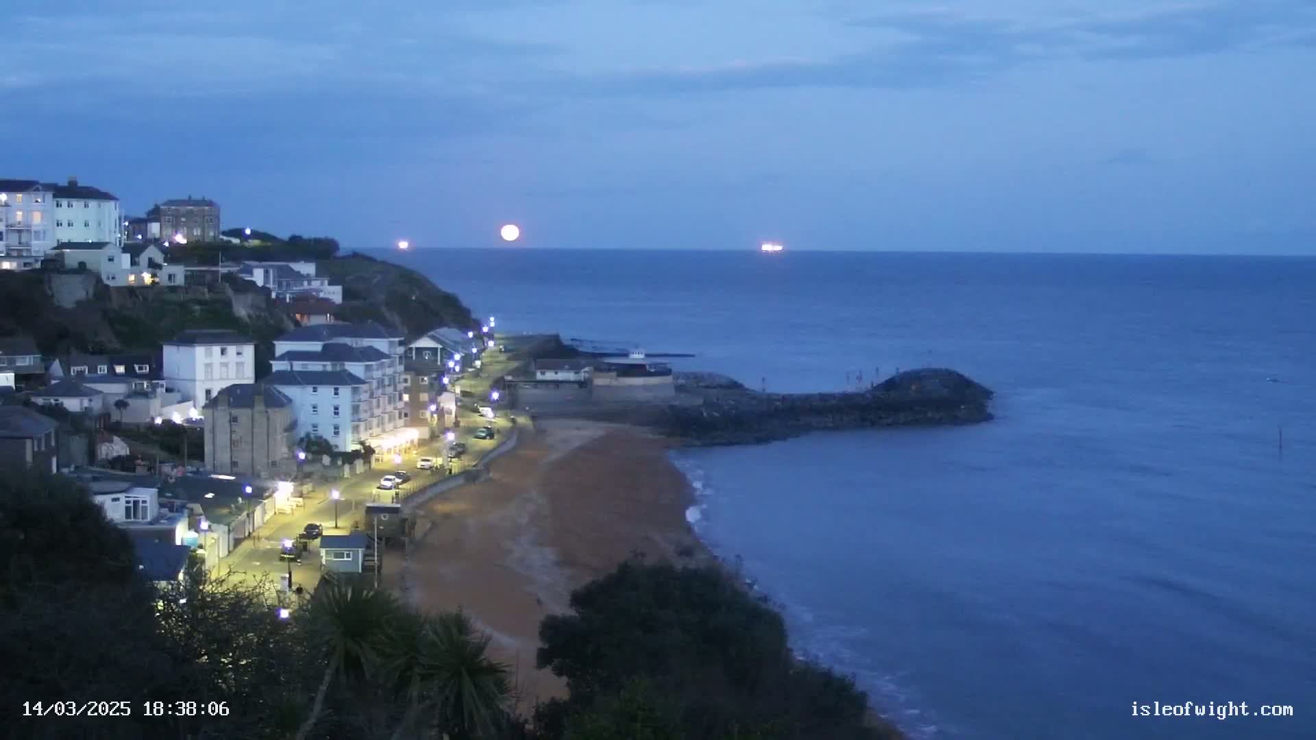 A coastal town at twilight, illuminated by streetlights, overlooks a calm, dark blue sea under a full moon.