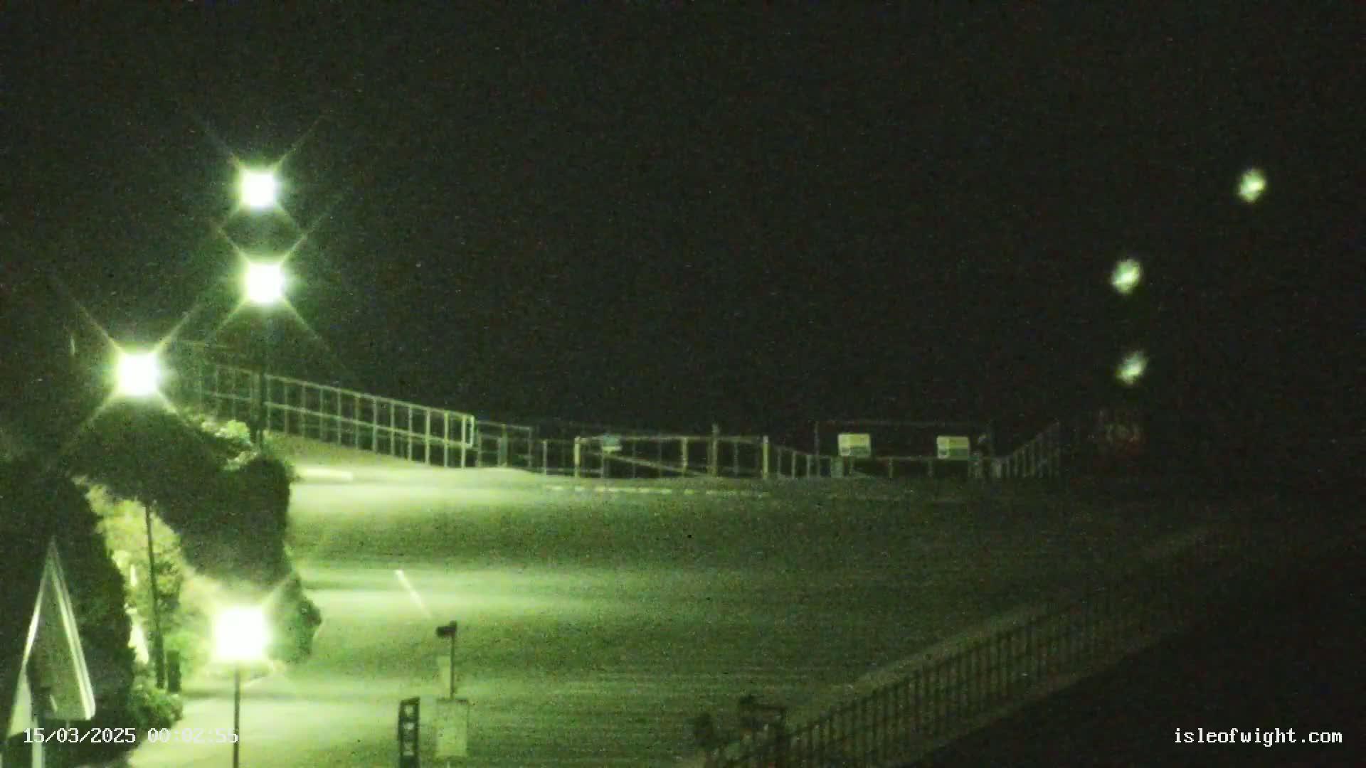 At night, several bright lights illuminate an empty sloped area with railings and sparse vegetation, under a dark sky.