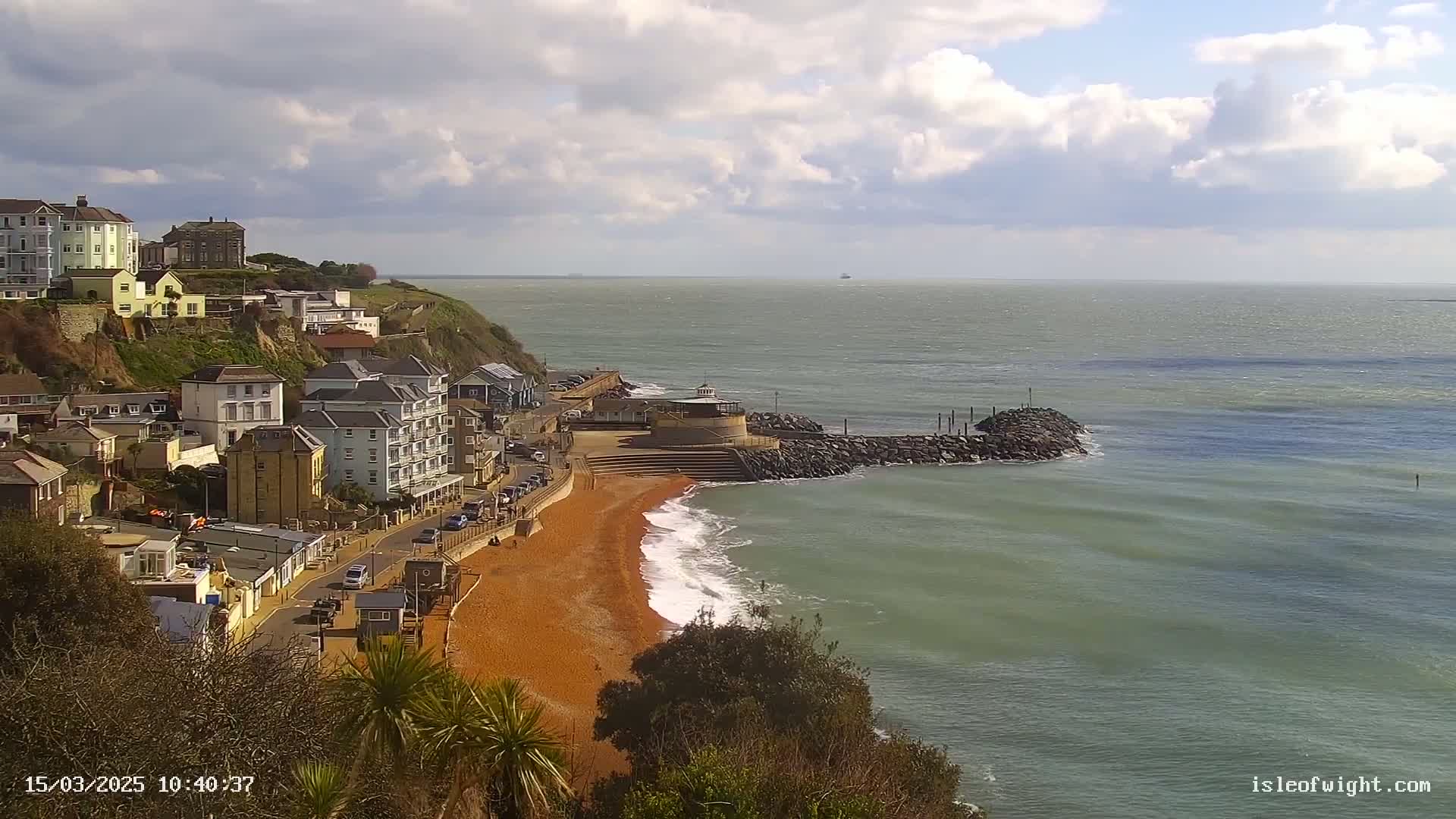 A red sand beach curves gently around a rocky breakwater, with buildings and houses perched on a cliff overlooking a calm sea under a partly cloudy sky.