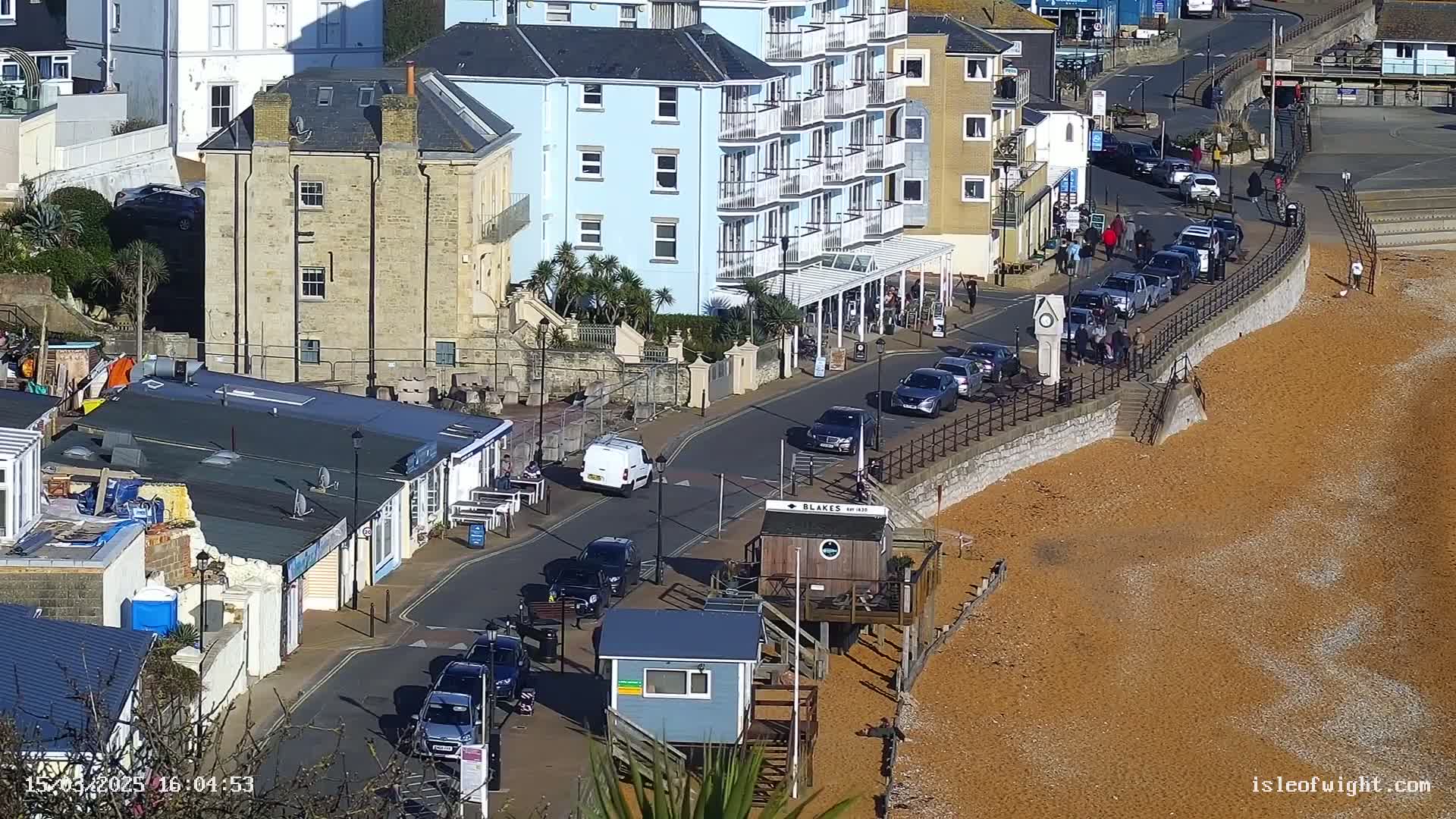 A sunny coastal town scene shows cars parked along a street next to a beach of reddish-brown shingle, with several buildings, including a multi-story light blue one, visible in the background.