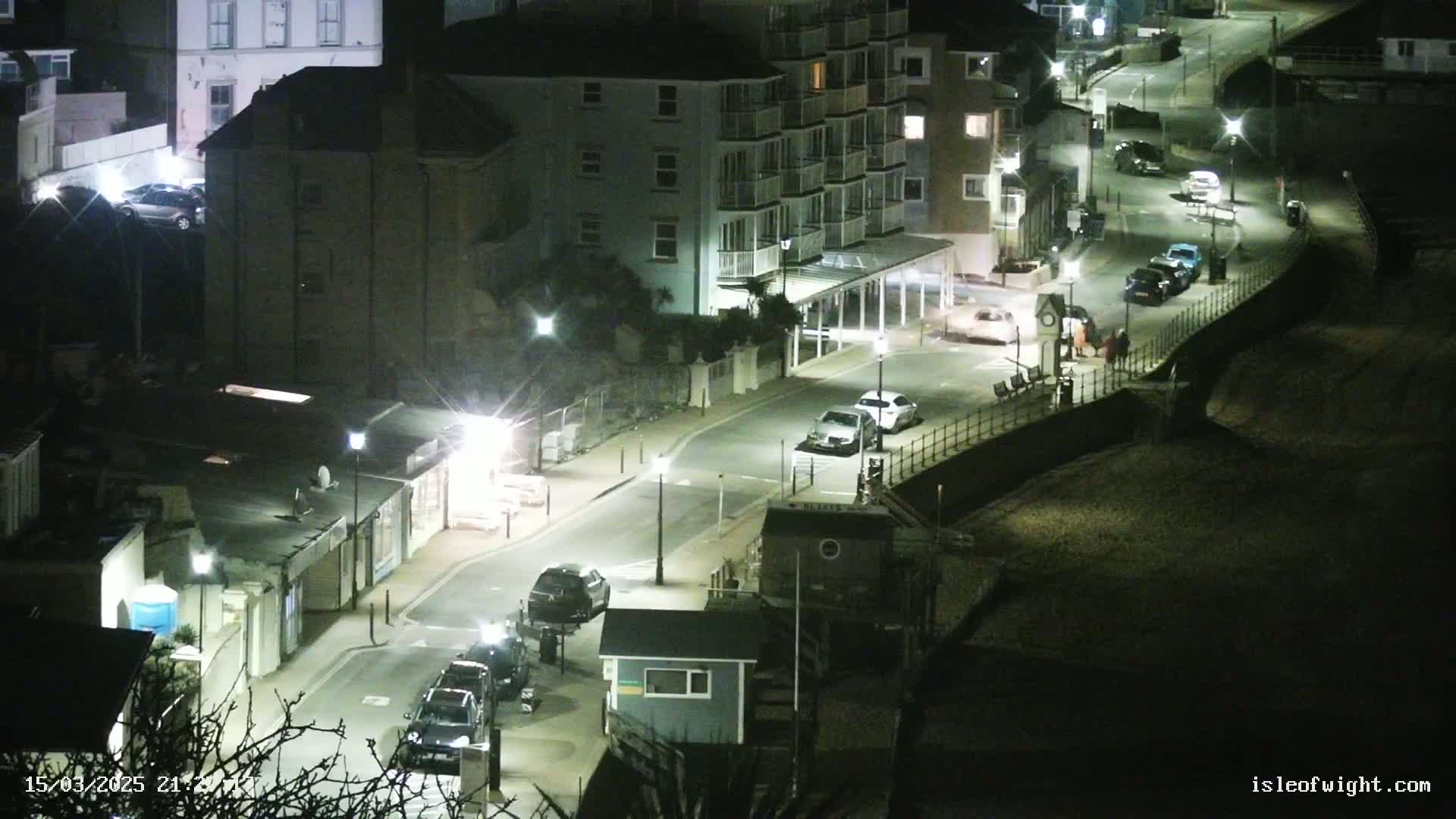 A nighttime aerial view of a seaside town street with parked cars and a few pedestrians, lit by streetlights.