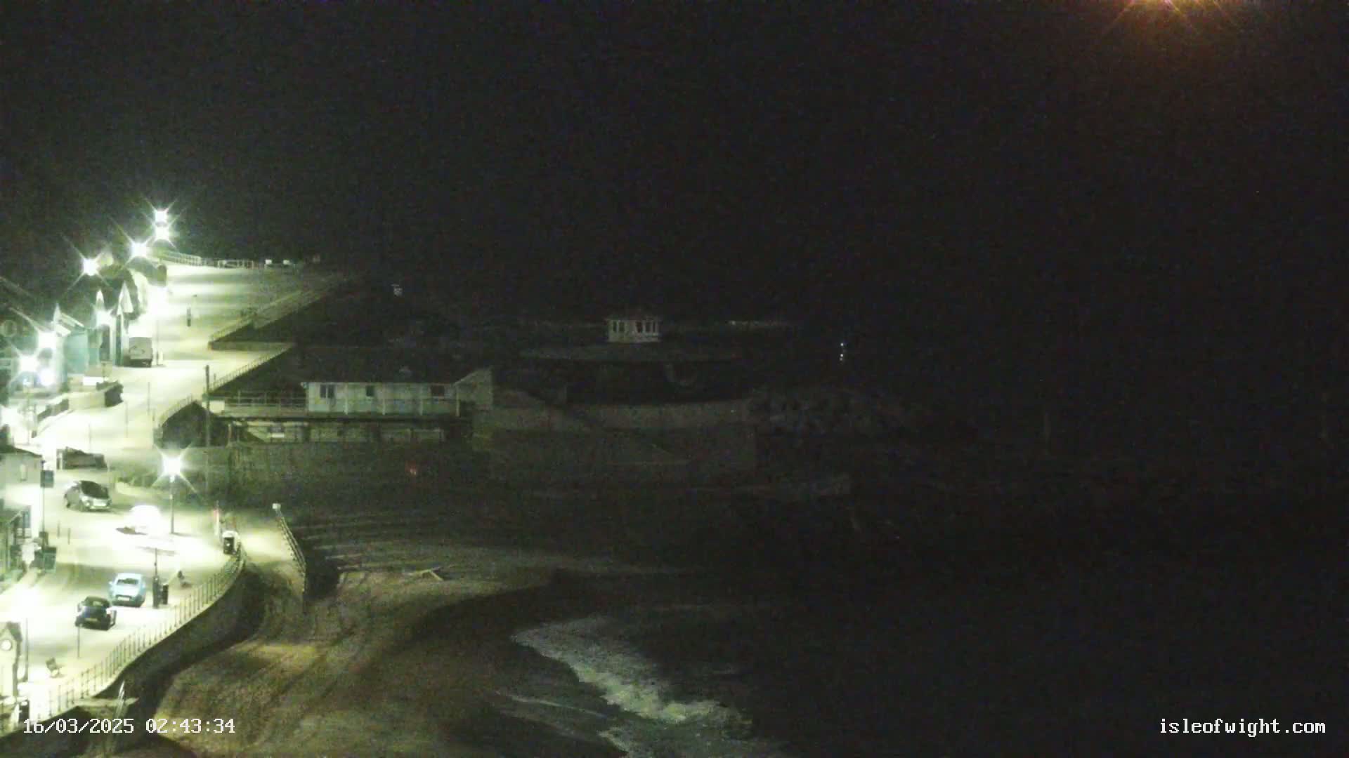 A nighttime coastal scene shows a dark beach with small waves, a curving lit roadway with parked cars, and buildings along the shore under a dark sky.