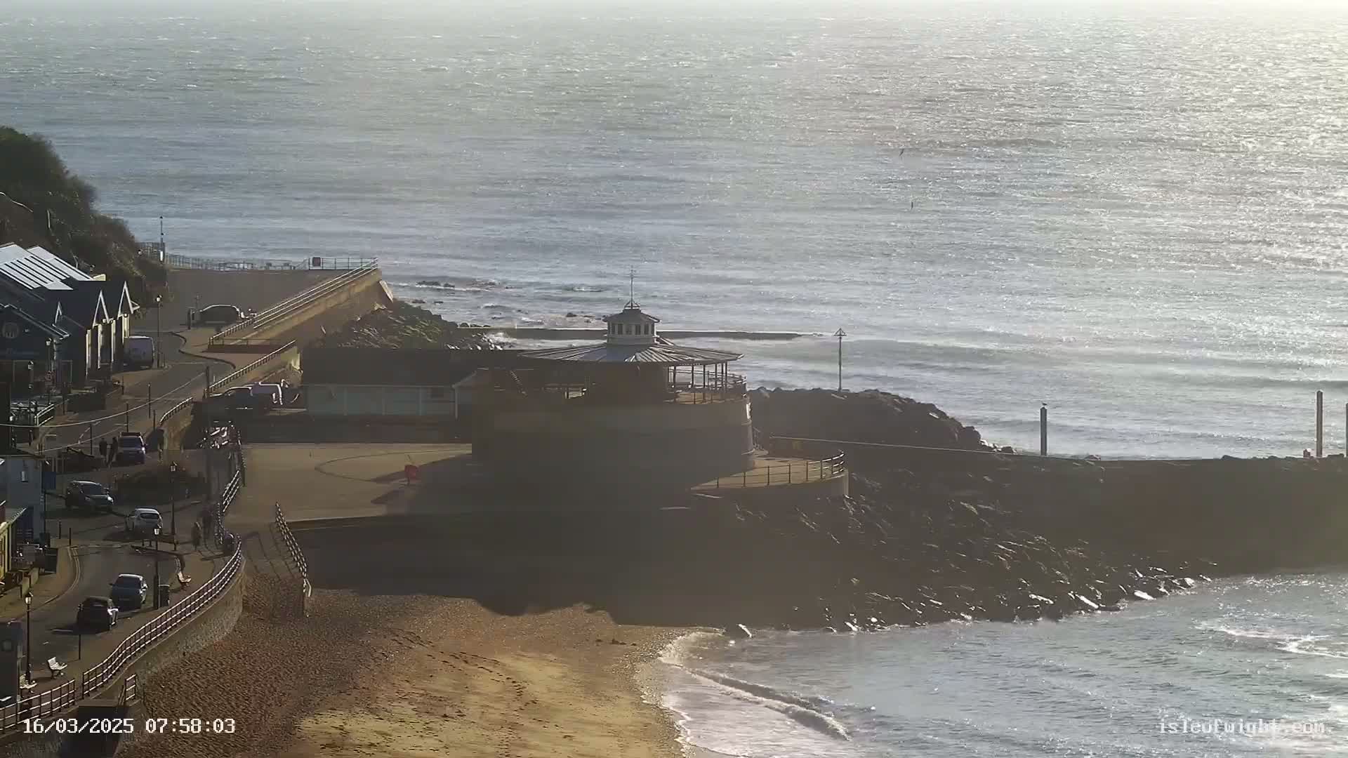 A seaside scene on a sunny day shows a sandy beach, a small building with a cupola on a rocky outcropping, a road with cars, and calm ocean waves.