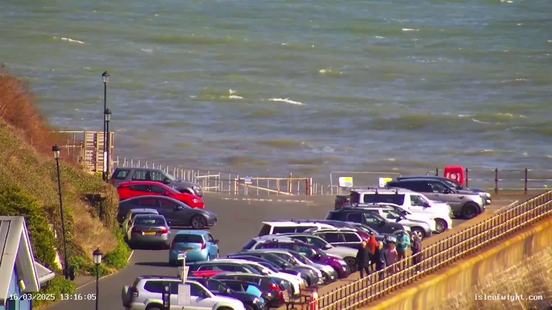 A parking lot filled with cars is situated on a cliff overlooking a choppy sea on a sunny day.