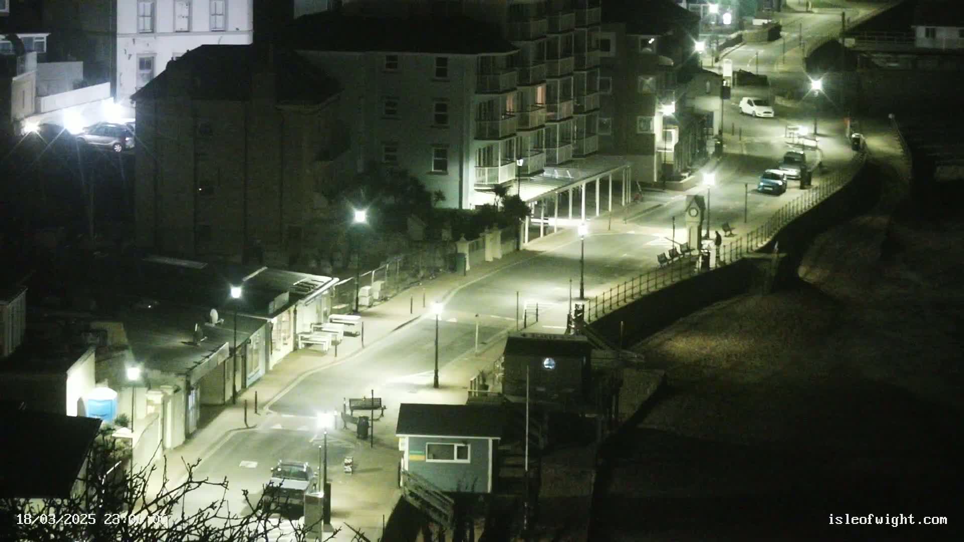 A nighttime, overhead view of a seaside street with buildings, parked cars, and streetlights illuminating the scene.