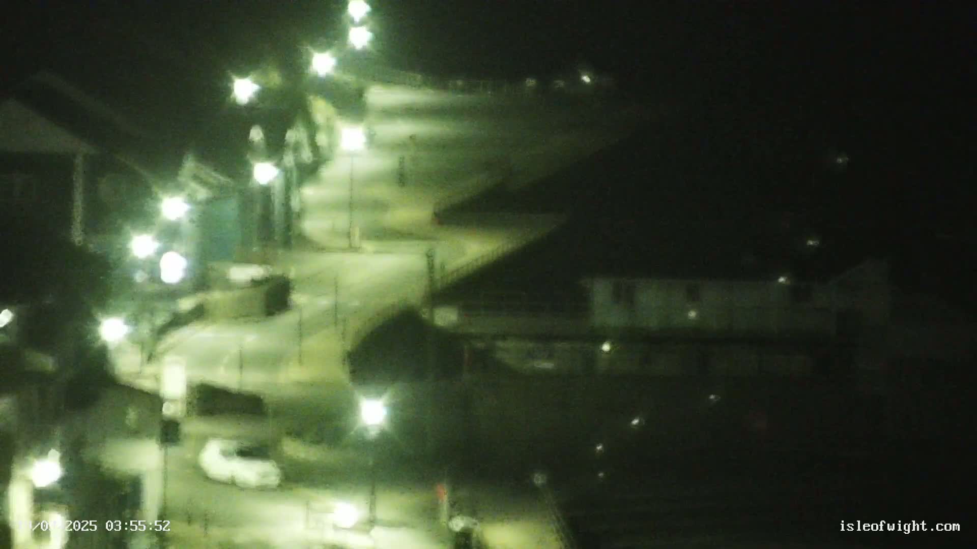 A nighttime, high-angle view shows a curving road illuminated by streetlights, with a few vehicles visible, and buildings flanking the road.