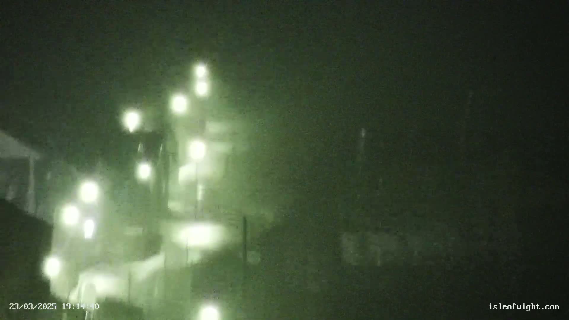 A blurry nighttime image shows a structure illuminated by multiple lights in hazy or foggy conditions.