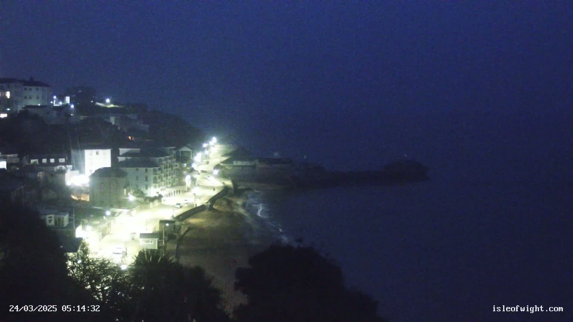 A dimly lit coastal town at night is shrouded in fog, with the lights of buildings and a street visible along a dark beach and ocean.