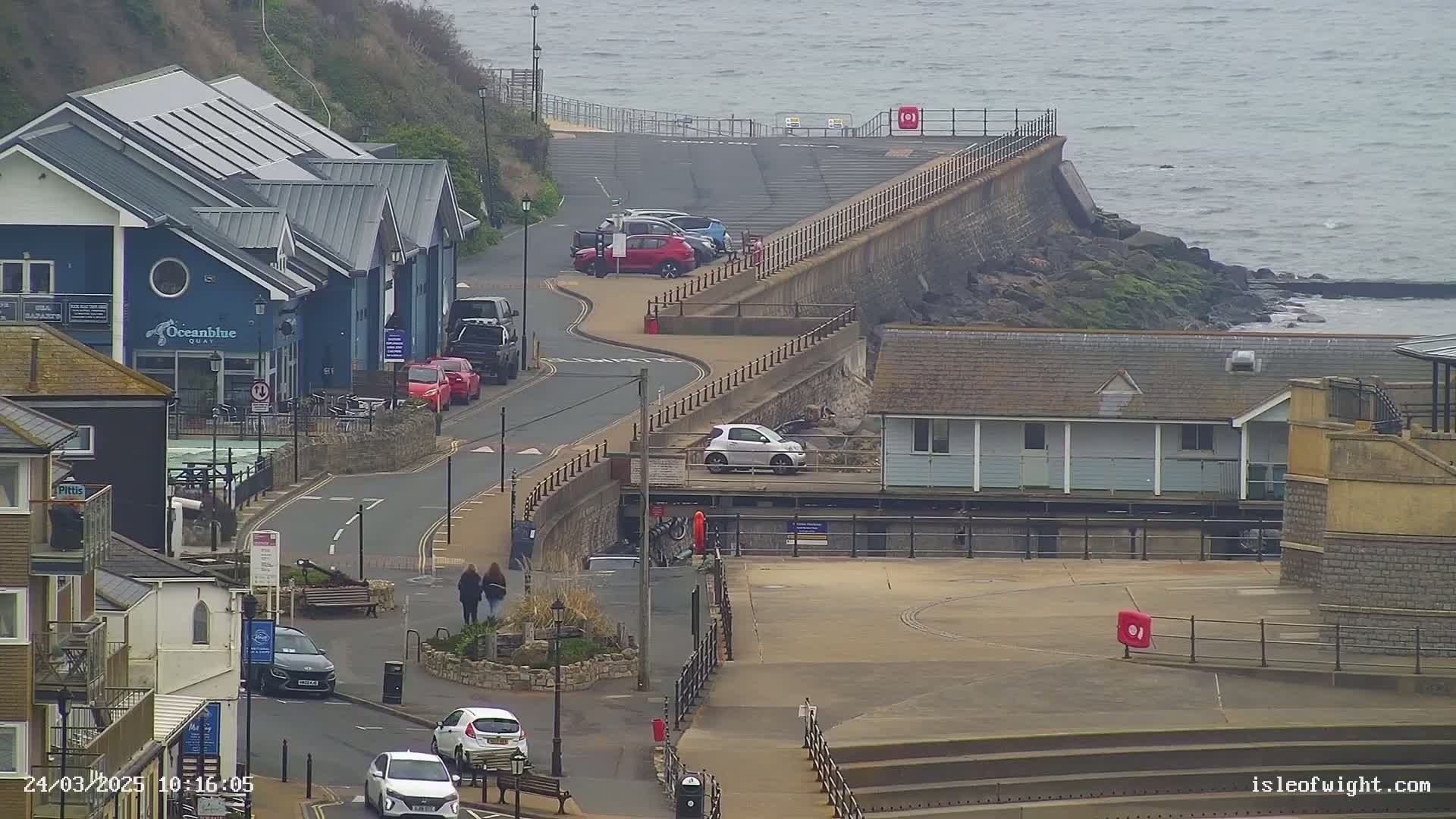 A seaside town scene on an overcast day shows a curving road with parked cars leading to a waterfront area with buildings and a seawall bordering the rocky shore.