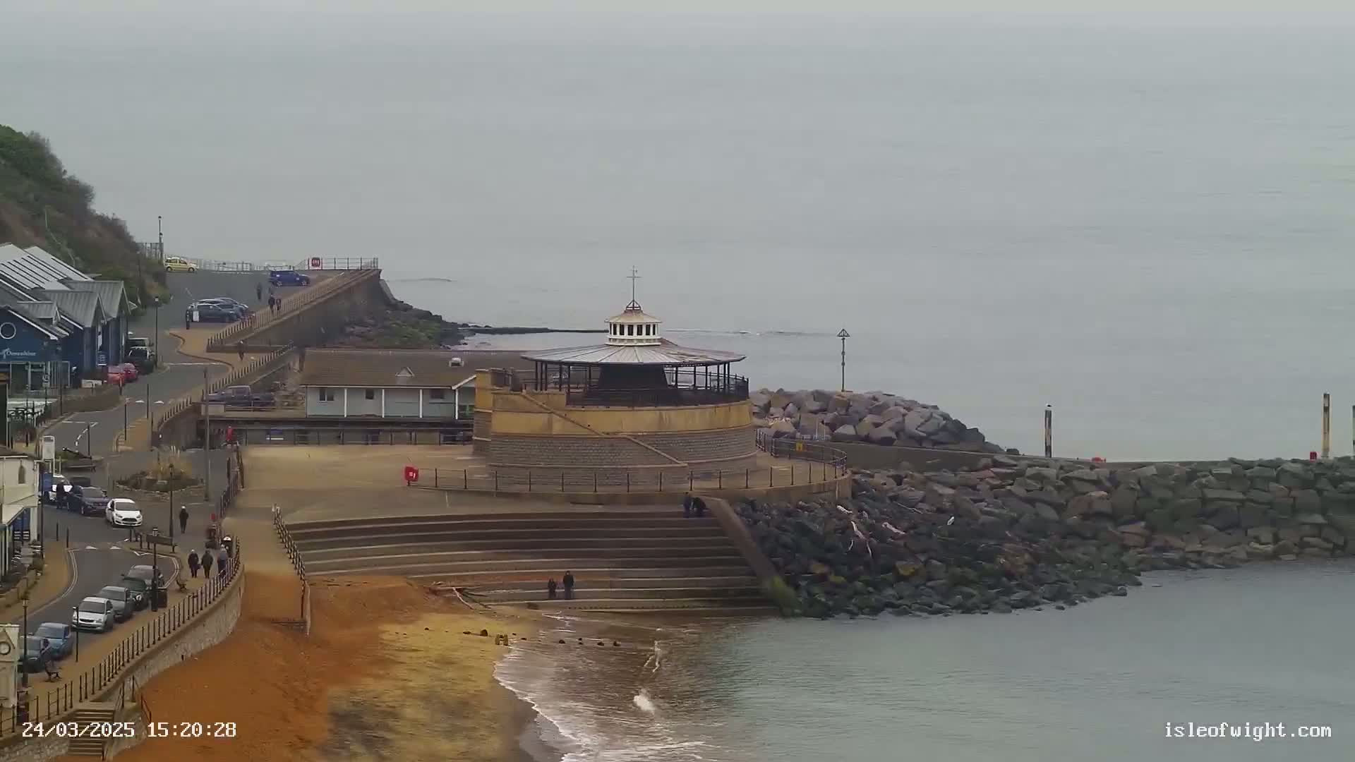 A seaside scene on an overcast day shows a circular bandstand on a stone structure overlooking a rocky beach and calm water.