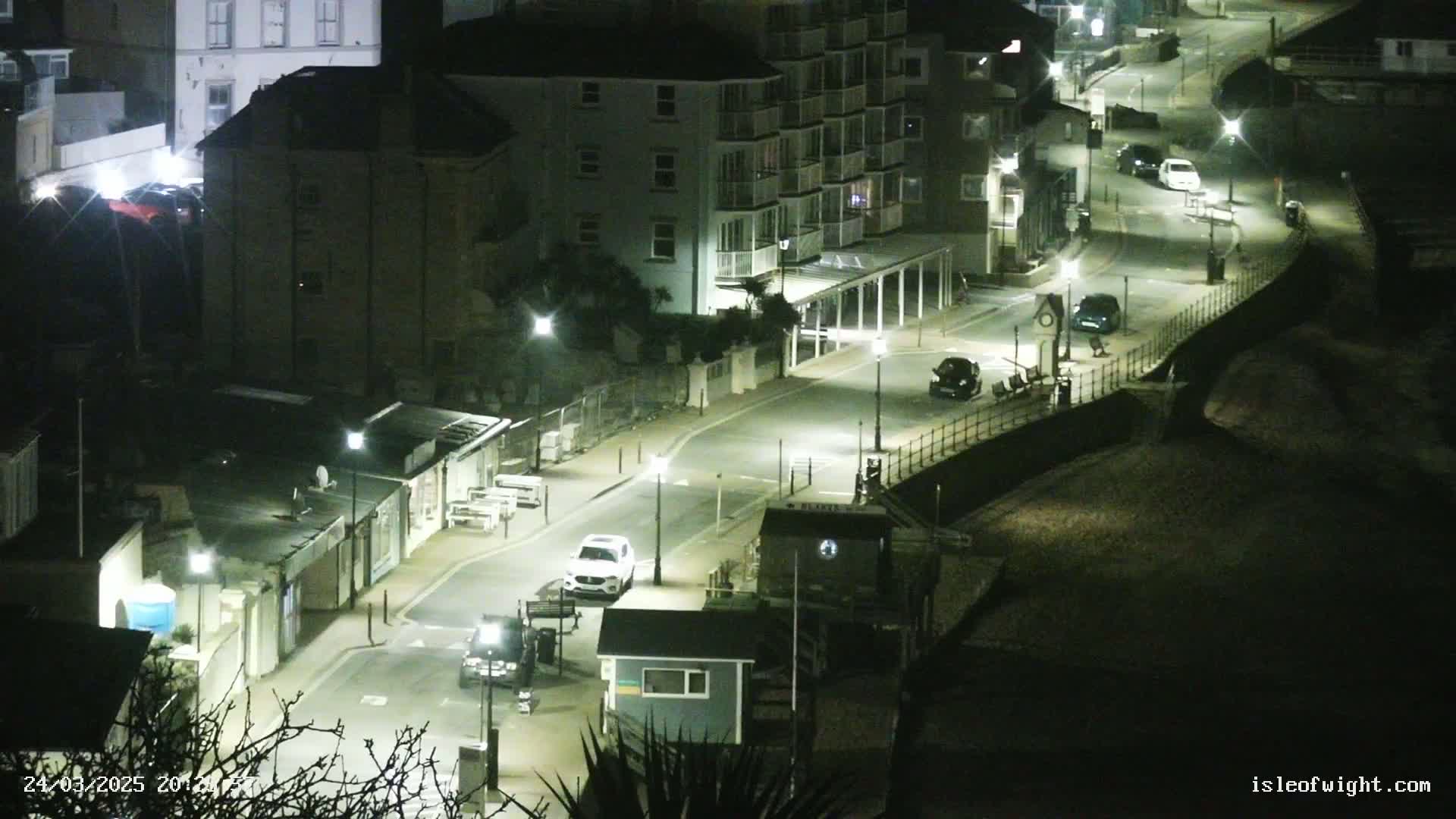A nighttime, high-angle view shows a curving street lined with buildings, streetlights, and a few parked cars.