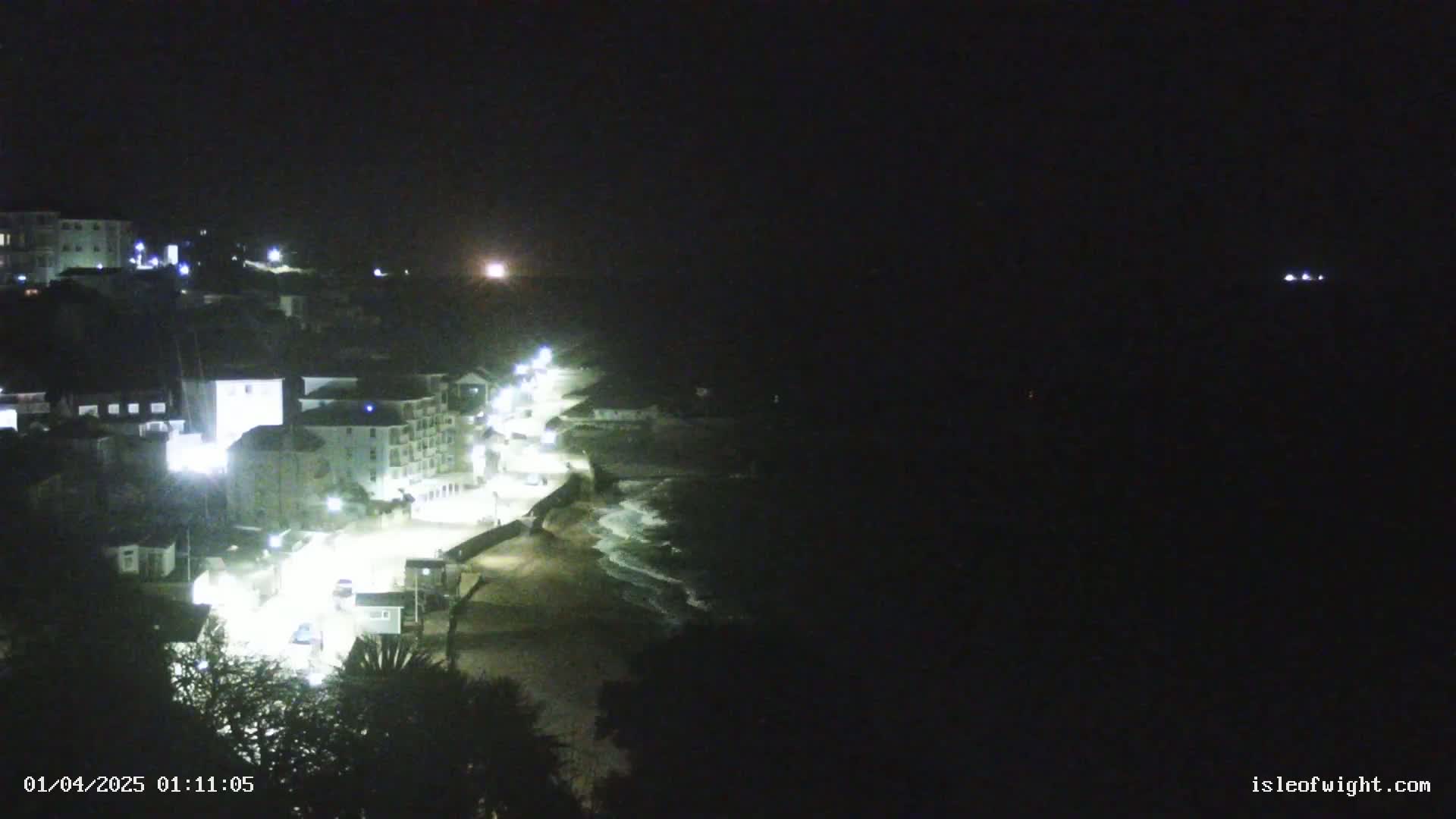 A nighttime coastal town with illuminated buildings and a dark ocean with waves gently lapping a sandy shore.
