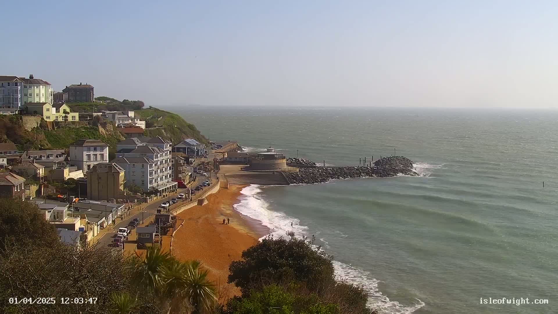 A seaside town with buildings on a cliff overlooking a sandy beach and calm sea under a clear sky.