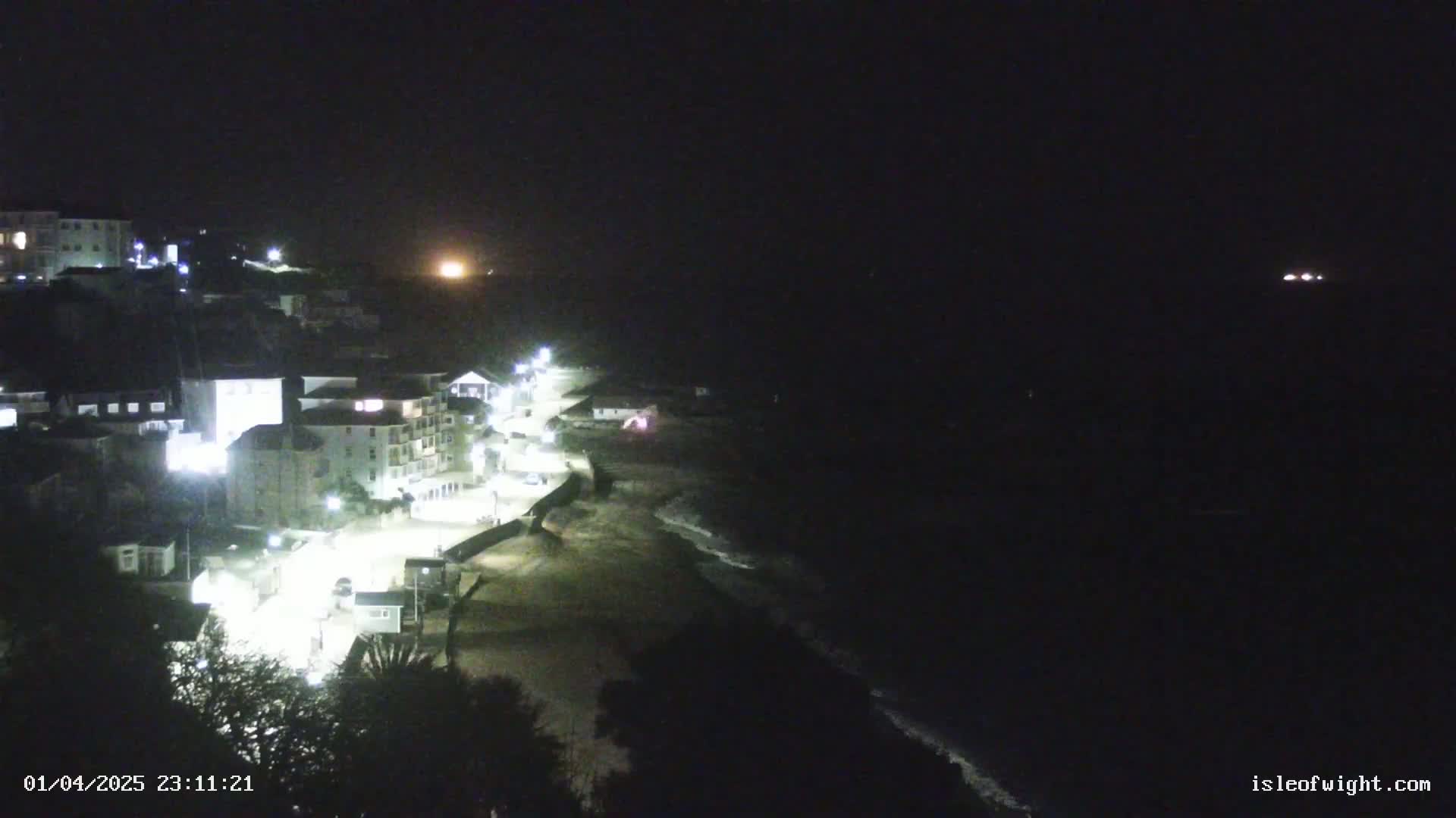 A nighttime coastal scene shows a dark, sandy beach, lit buildings, and a dark ocean under a clear sky.