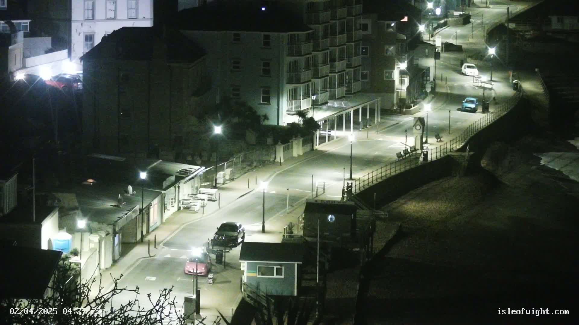 A nighttime, overhead view of a seaside street with several cars parked and driving, buildings and streetlights illuminating the scene.