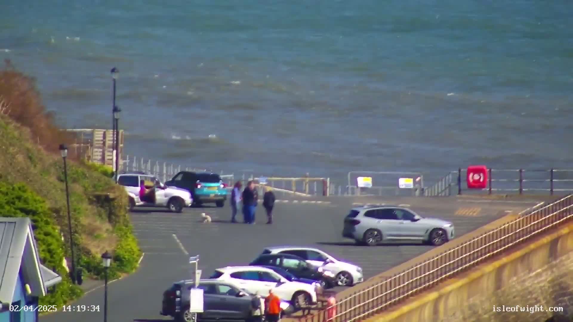 A parking lot with several cars overlooks a choppy sea on a sunny day.