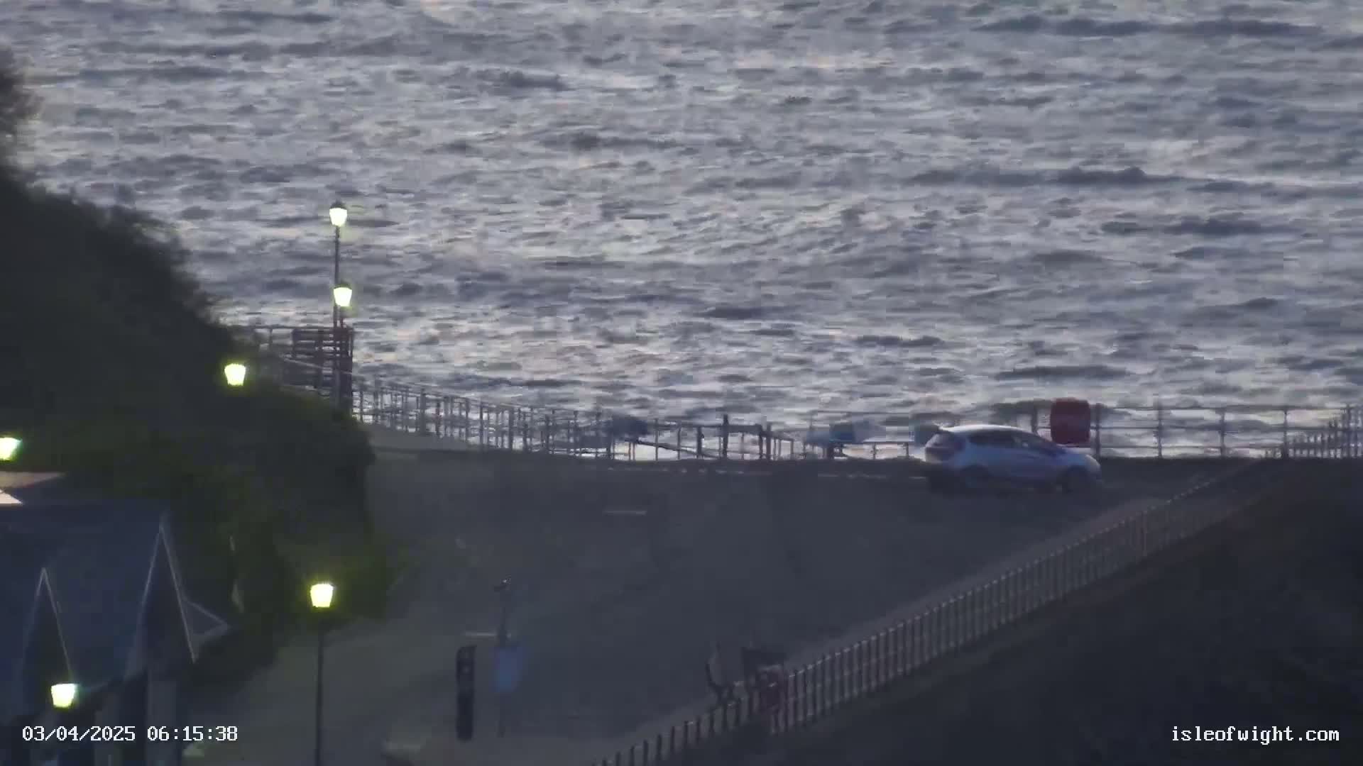 A car is parked near a fence overlooking a choppy sea under a dark, overcast sky.