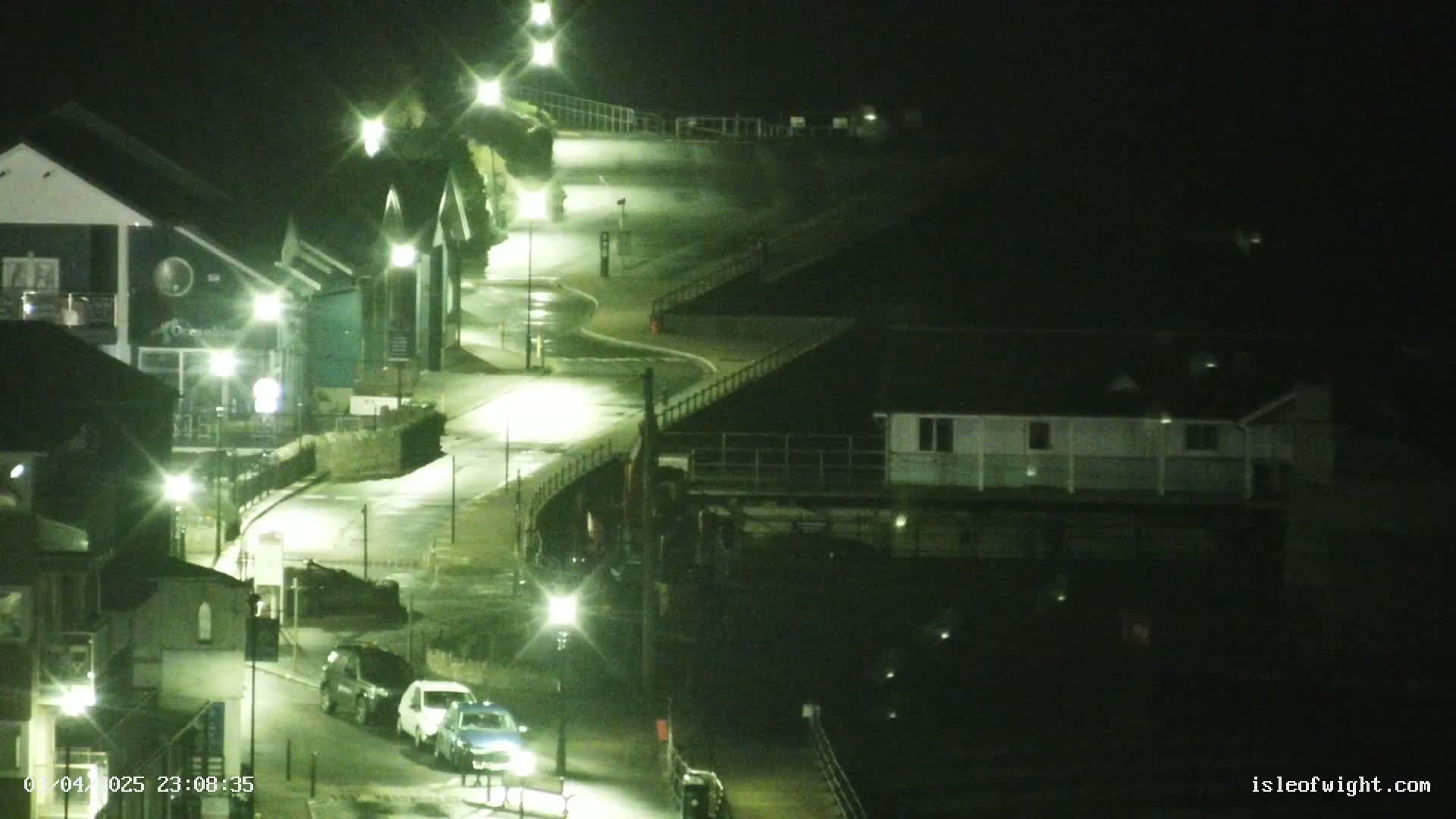 A rain-slicked, curving road at night, lit by streetlights, winds past buildings and a few parked cars.