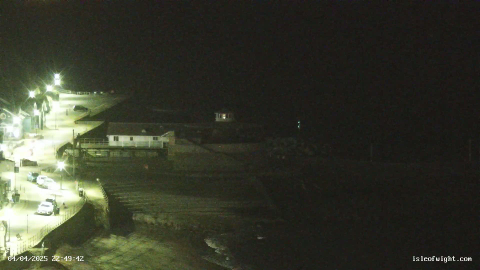 A nighttime coastal scene shows a road with parked cars curving along a seawall toward a small building, with a dark beach and ocean below.