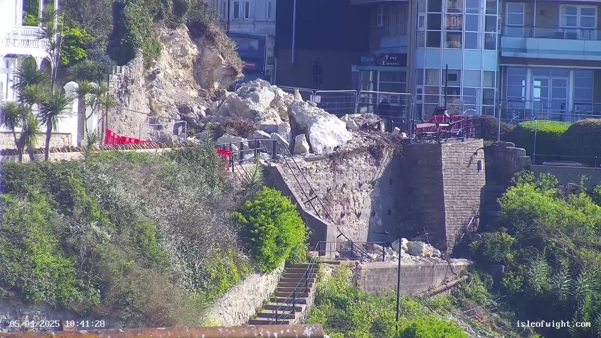 A large pile of rocks has fallen onto a stairway leading down a cliffside, near buildings on a sunny day.