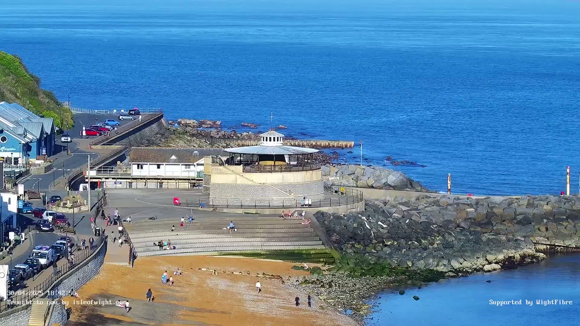A beach with a round gazebo and people is nestled between a rocky shoreline and calm blue ocean under a sunny sky.