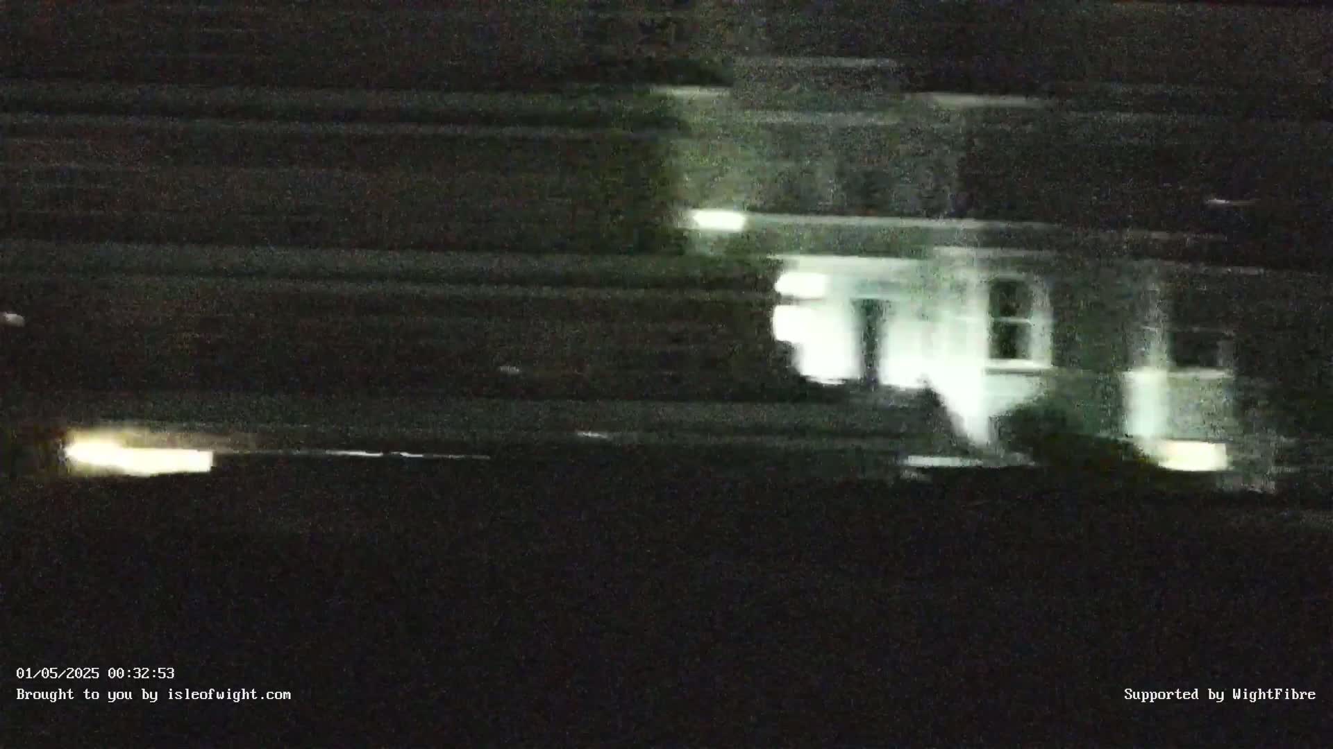 A blurry nighttime image shows a light-colored building and a vehicle's headlights in the distance.