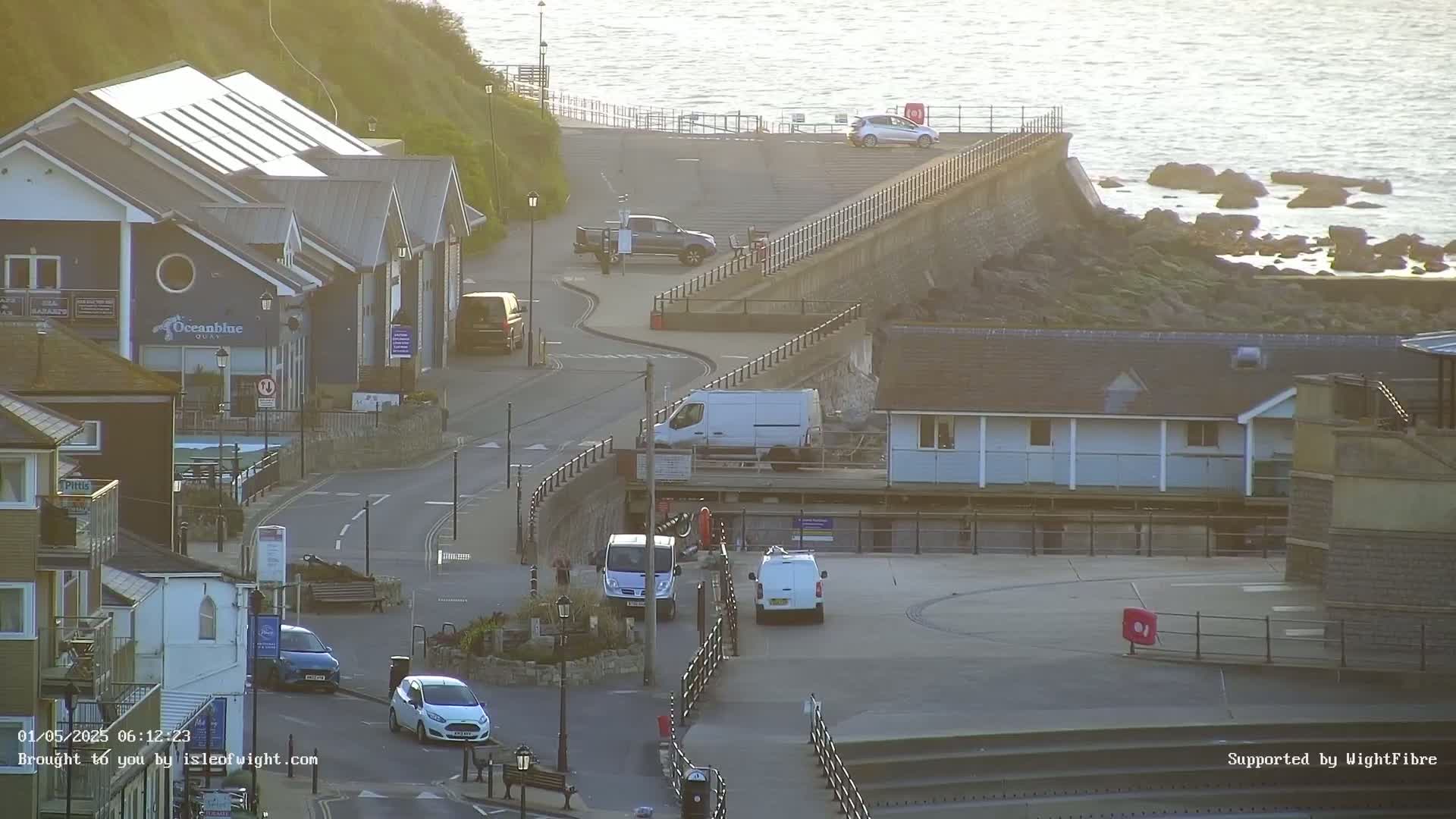A coastal town scene is shown on a sunny day, with several buildings, vehicles, and a sea wall overlooking a rocky shoreline and calm water.