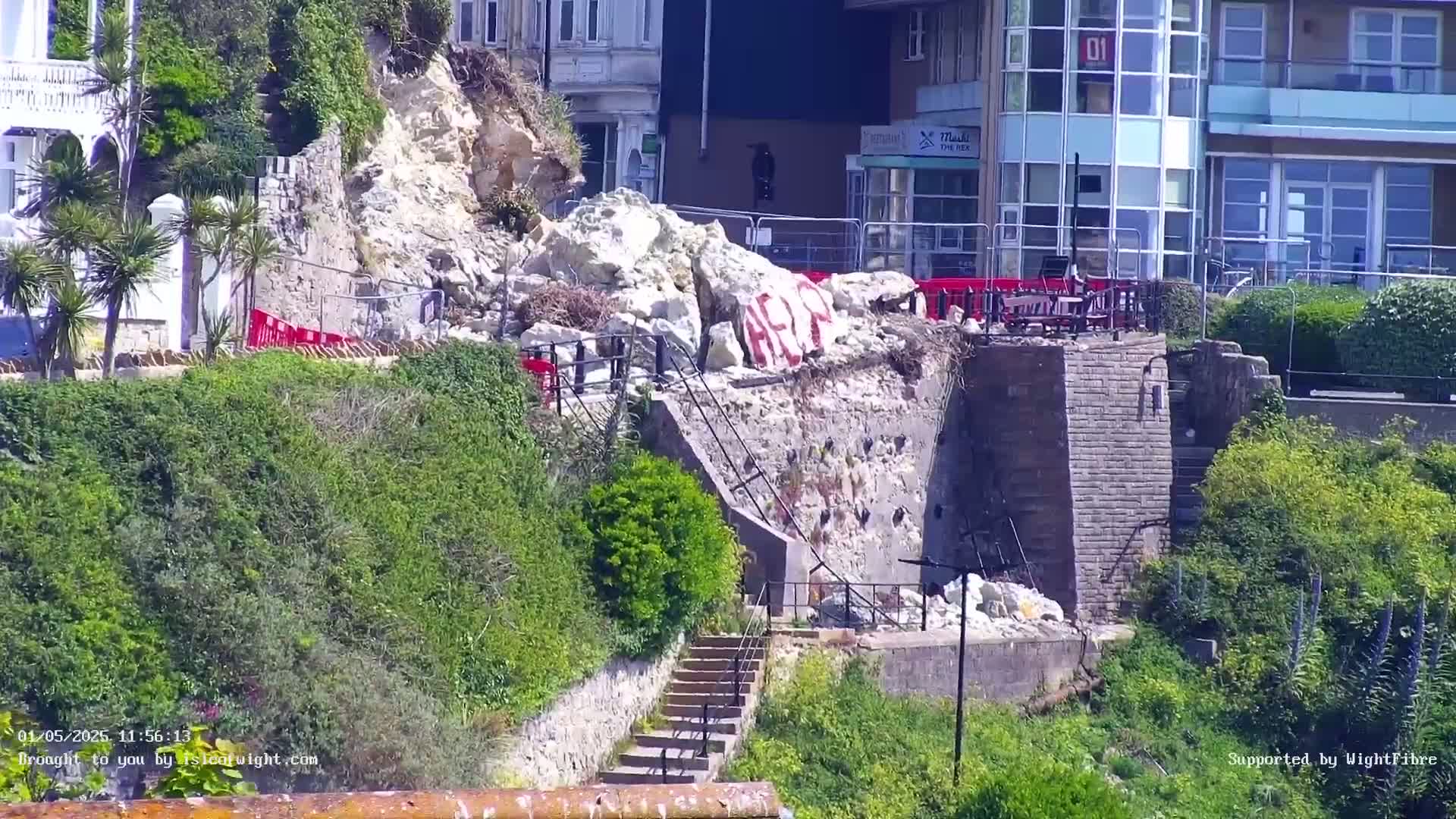 A large pile of rubble lies at the base of a stone wall next to a set of stairs, partially obscured by vegetation on a sunny day.