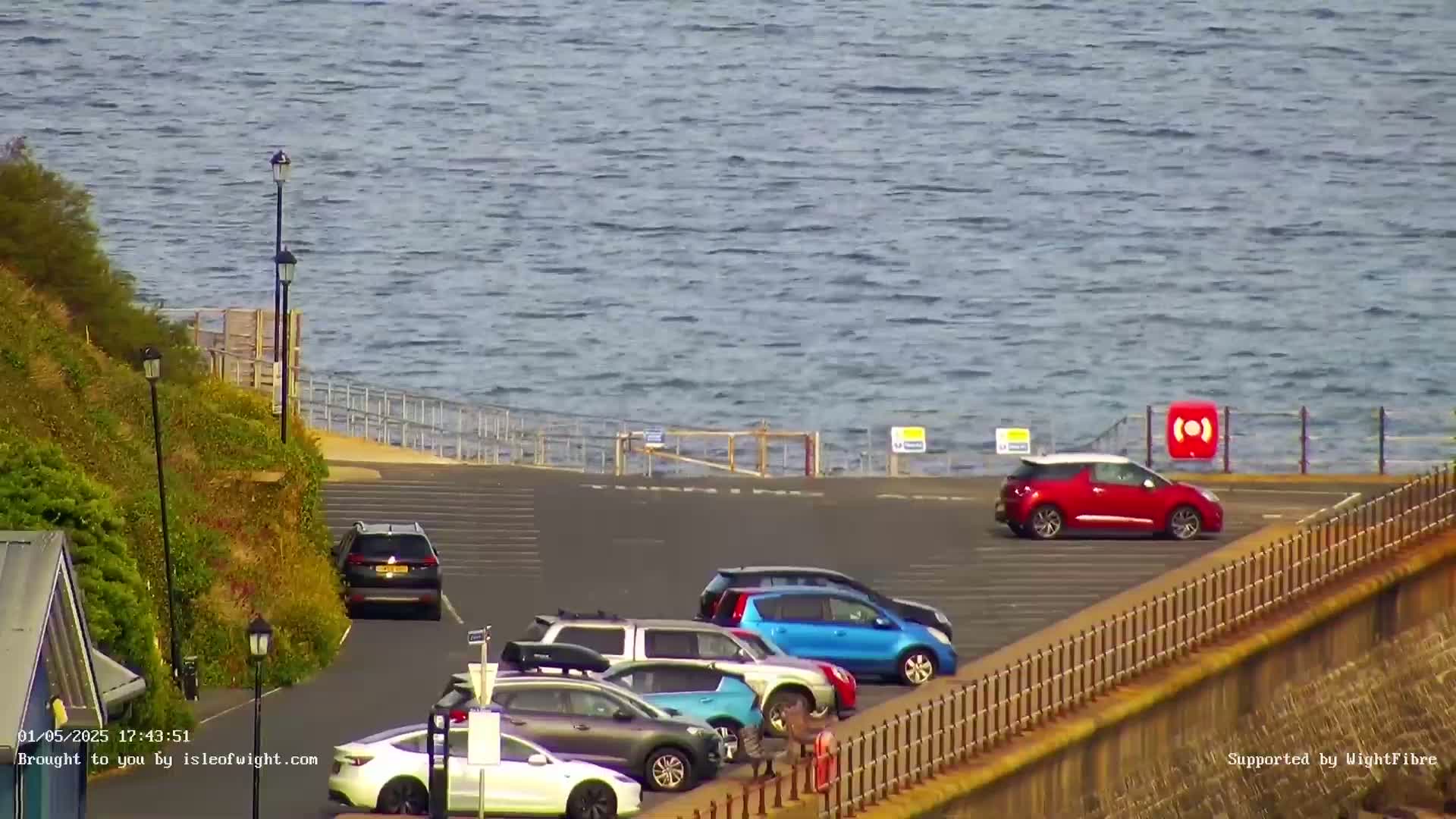 Several cars are parked in a lot overlooking a calm body of water on a sunny day.