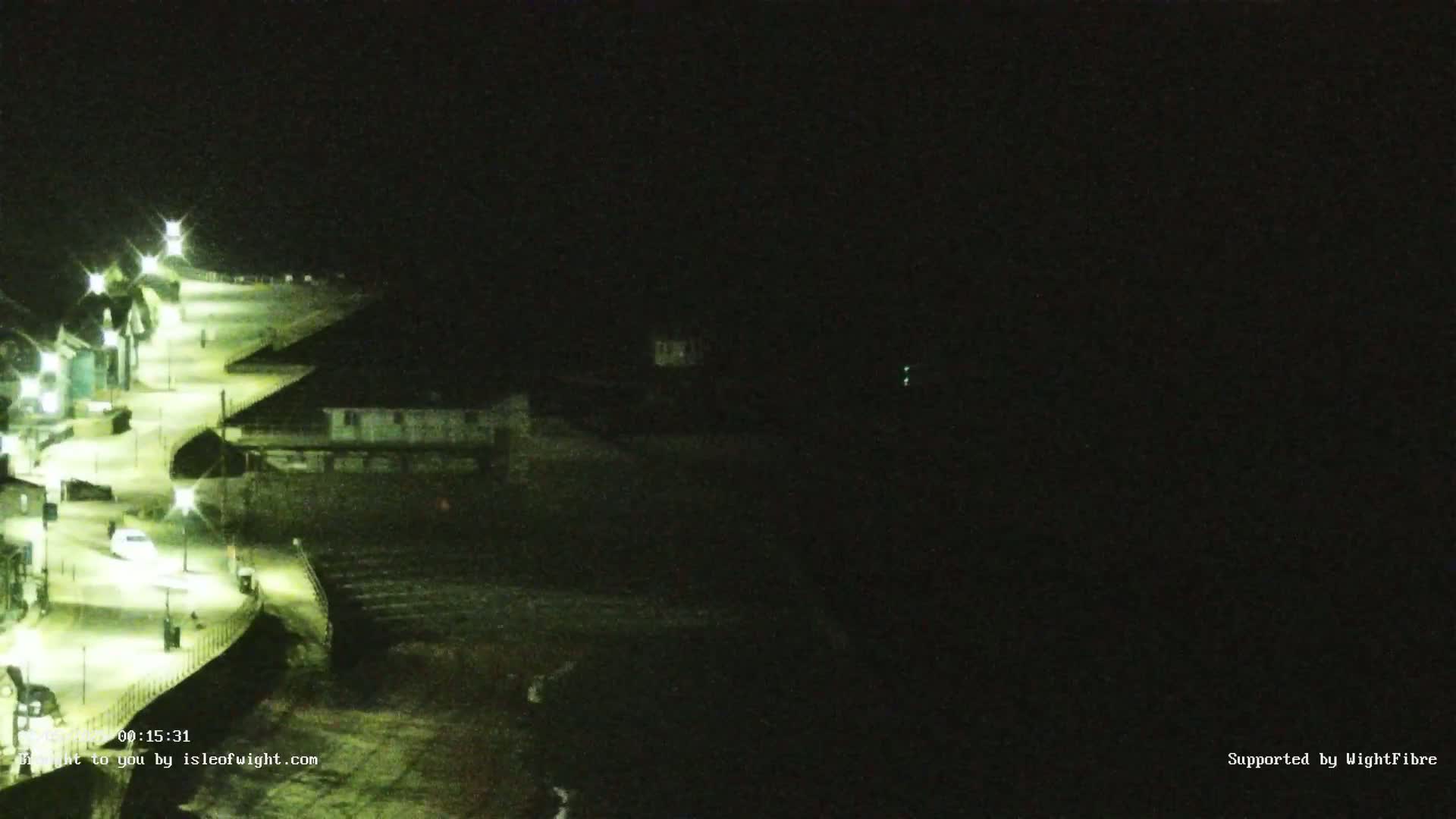 A nighttime, low-light view shows a paved walkway lit by streetlights curving along a dark shoreline.