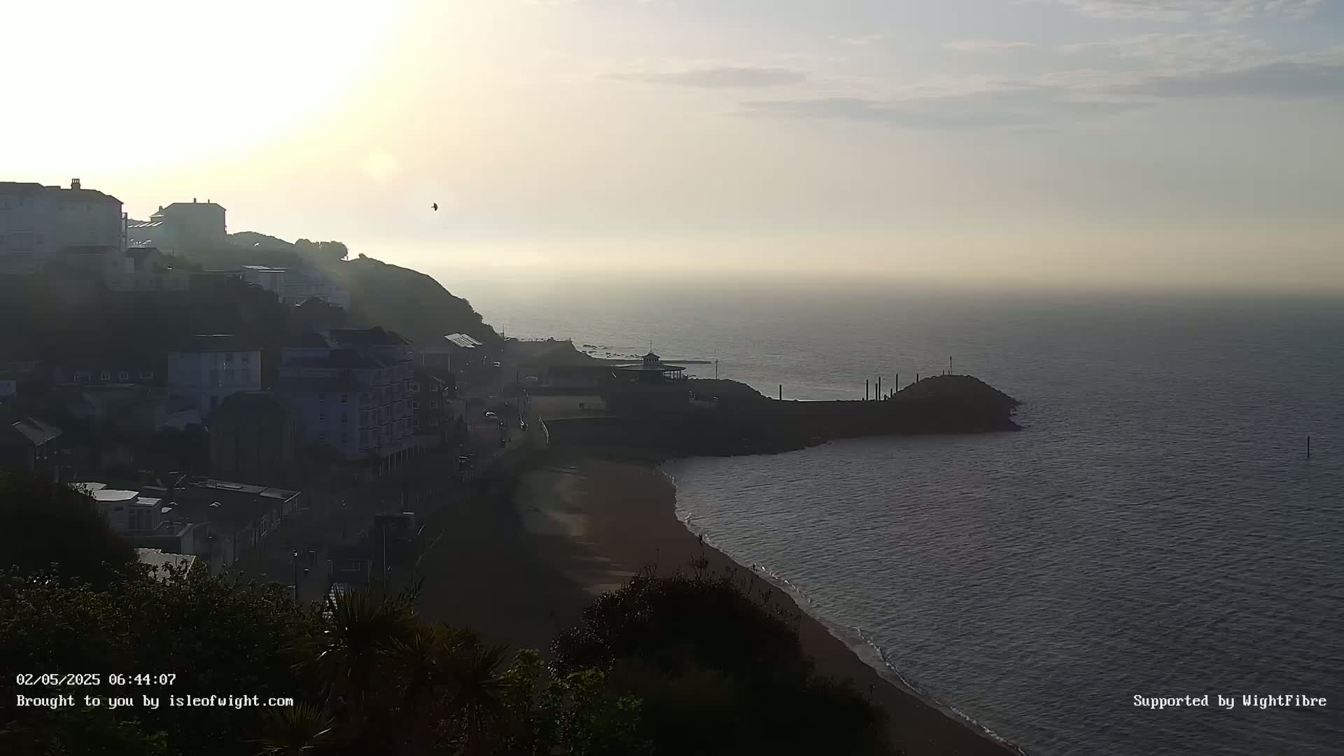 A hazy sunrise illuminates a coastal town nestled on a hillside overlooking a calm sea and sandy beach.