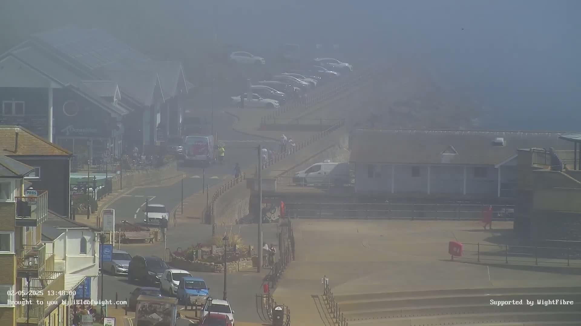 A foggy coastal town scene shows buildings, parked cars, pedestrians, and a road, with some vehicles in motion.