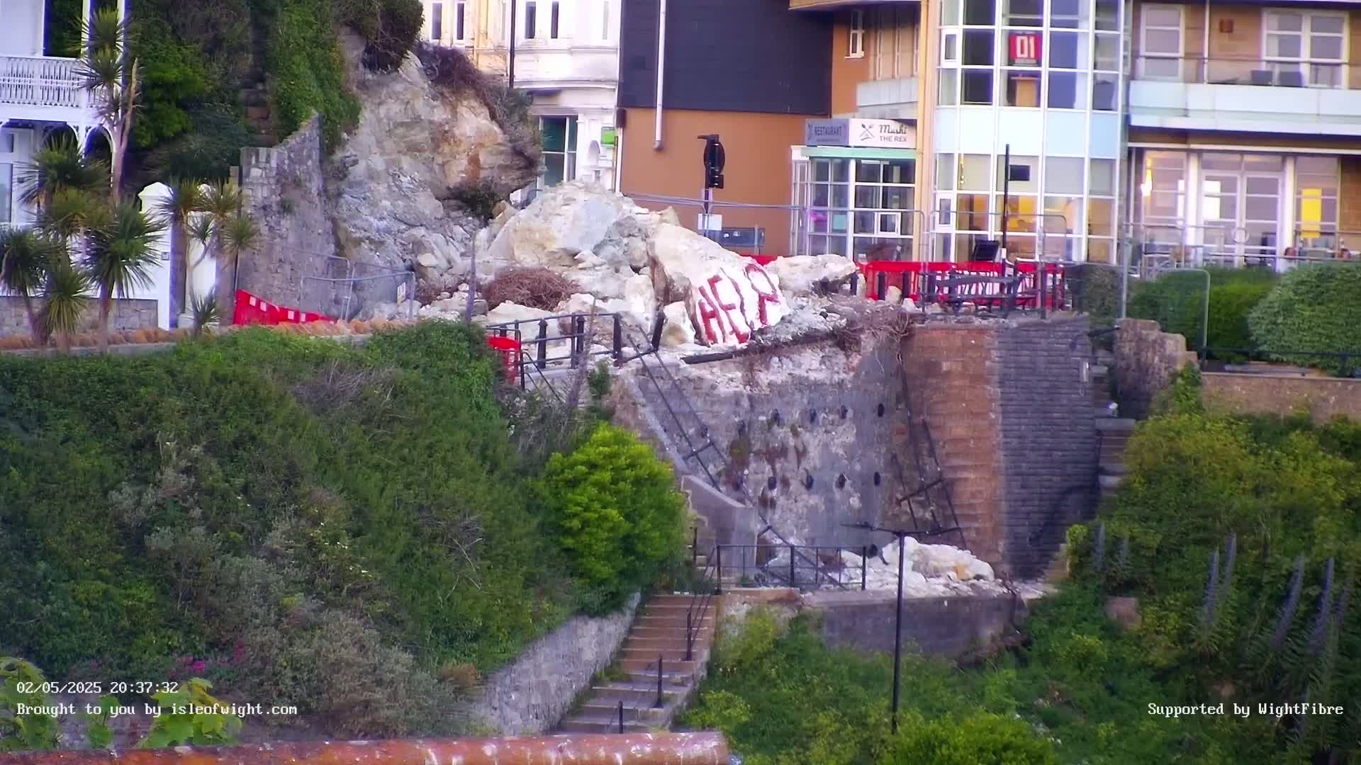 A section of a retaining wall has collapsed near a building, with large rocks and debris scattered below, and the word "HELP" spray-painted on a rock.